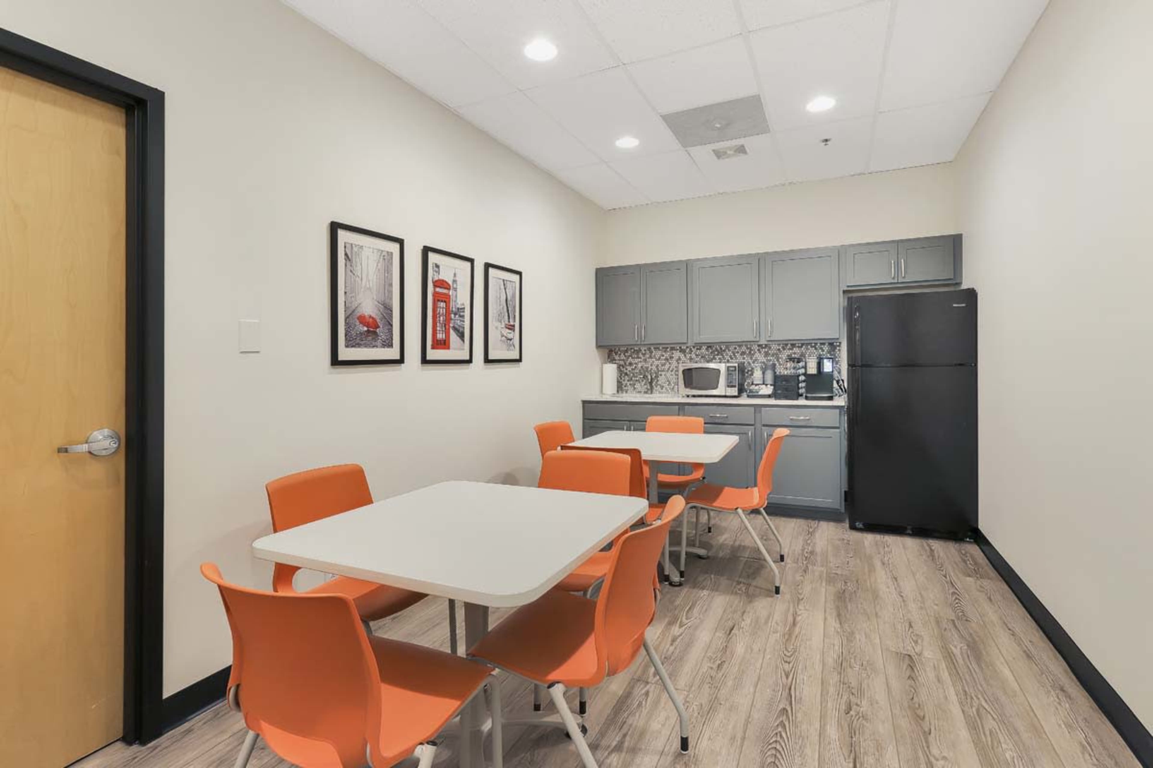 The image shows a modern break room with orange chairs, white tables, and a kitchenette featuring a microwave and black refrigerator.
