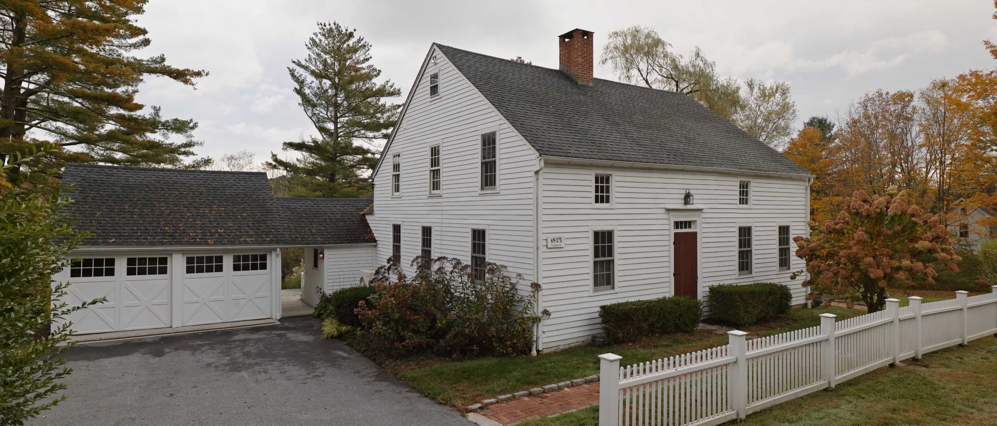 The image shows a two-story white house with a dark roof, a brick chimney, and a spacious front yard surrounded by a white picket fence.
