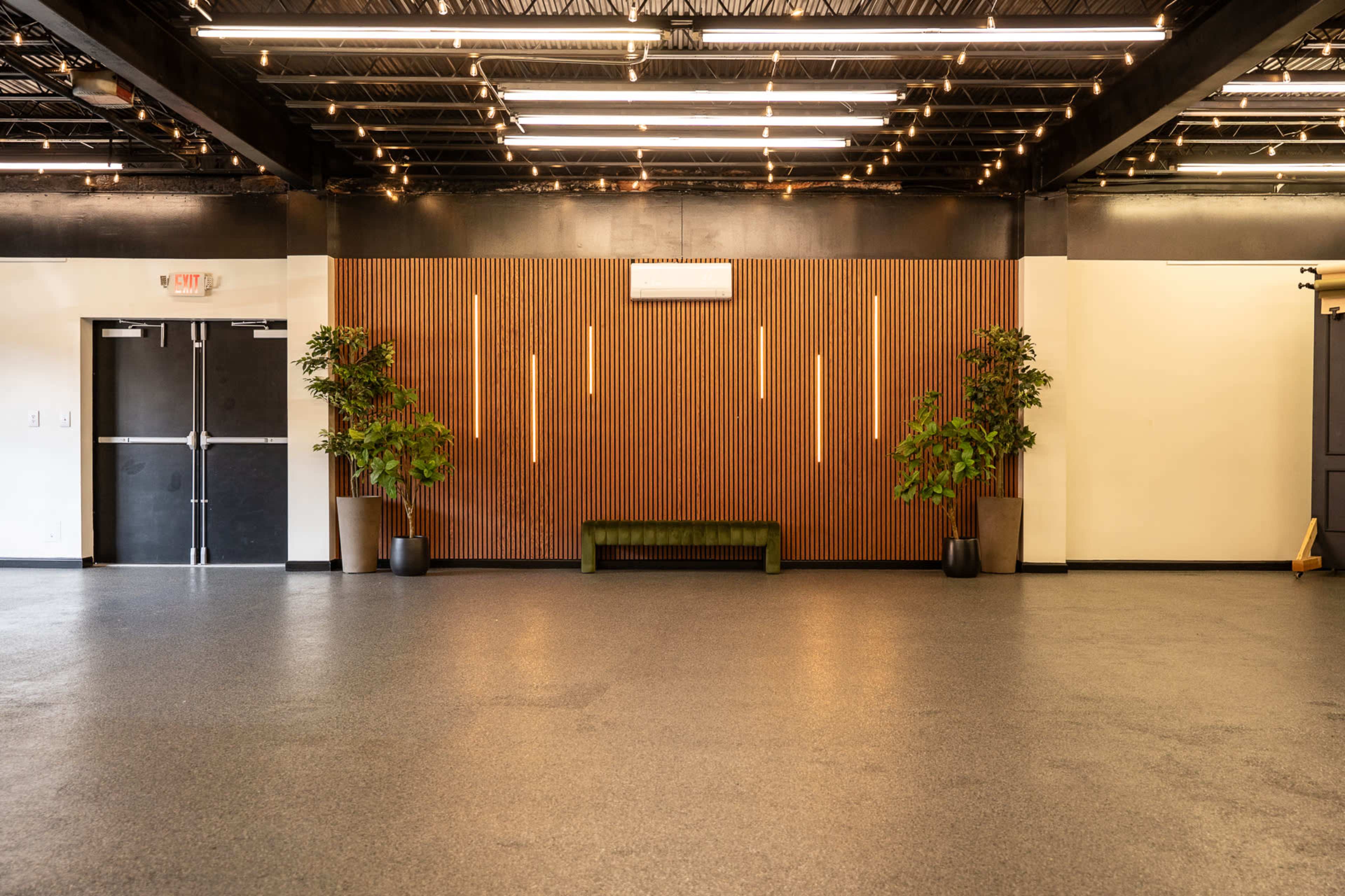 The image shows an empty interior space with a dark floor, wooden wall paneling, a green bench, and potted plants on either side.