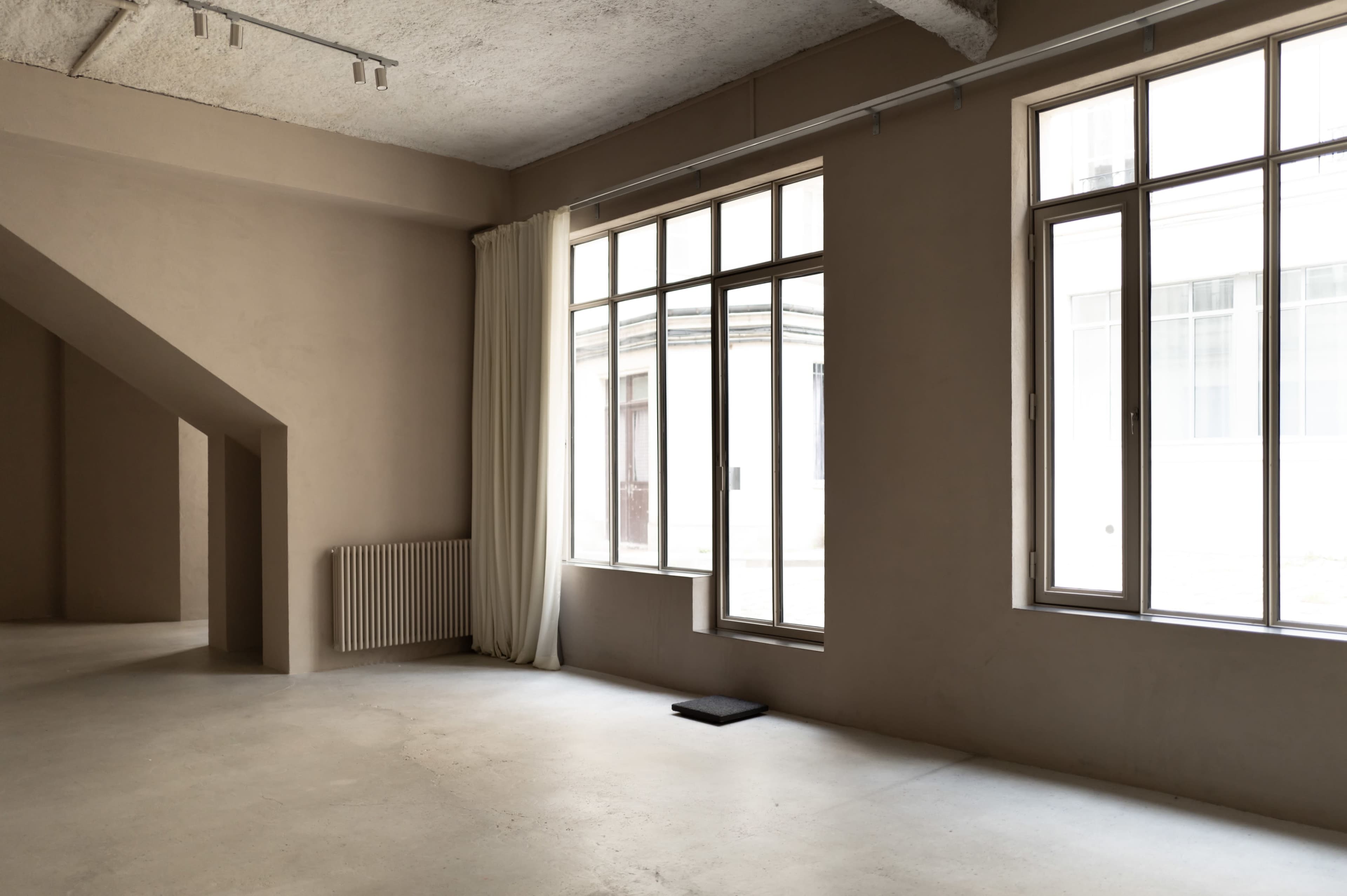 An empty interior space features large windows, light-colored walls, and a radiator along one wall.