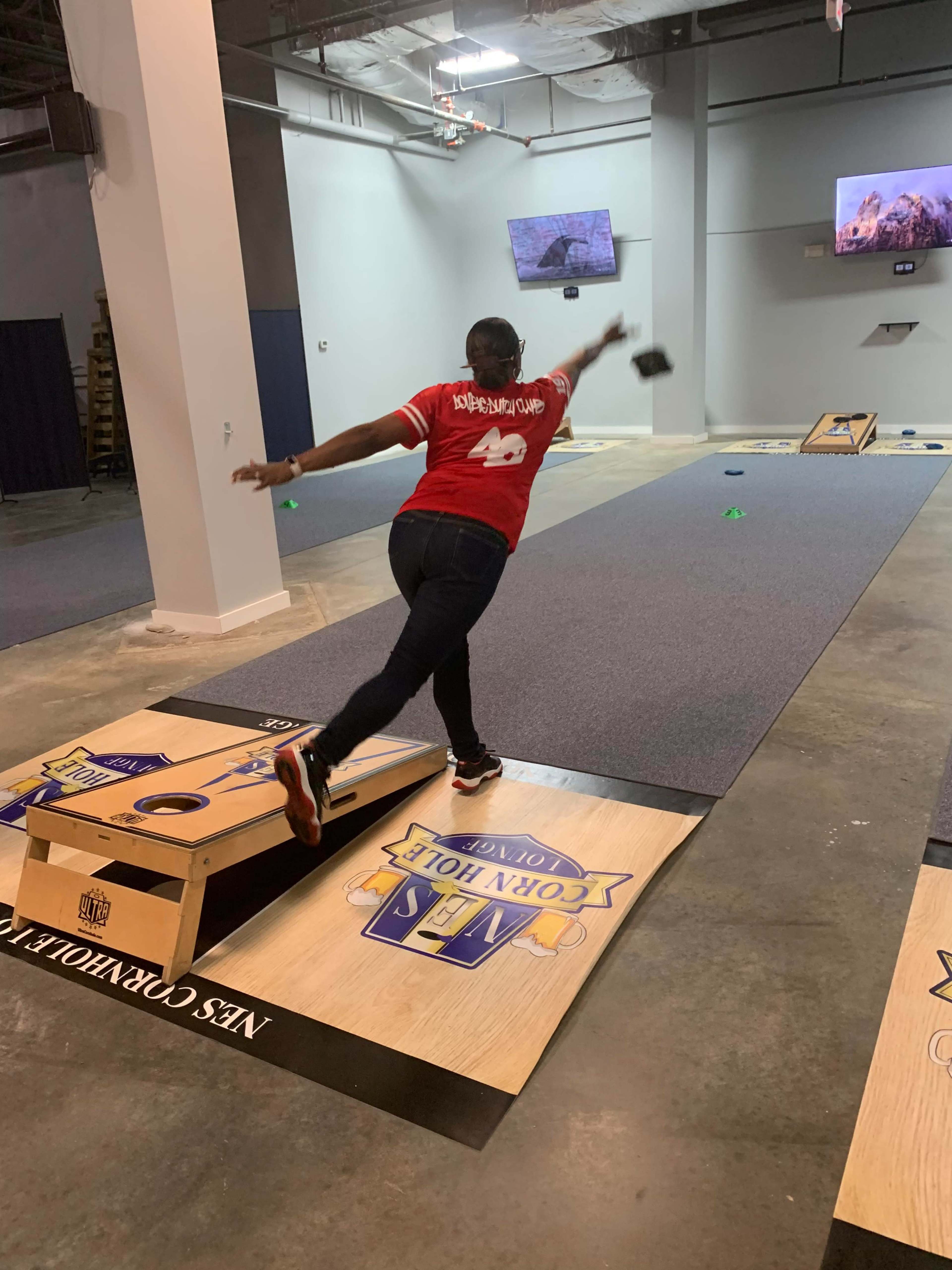 A person in a red shirt is throwing a beanbag towards a cornhole board in an indoor recreation area.