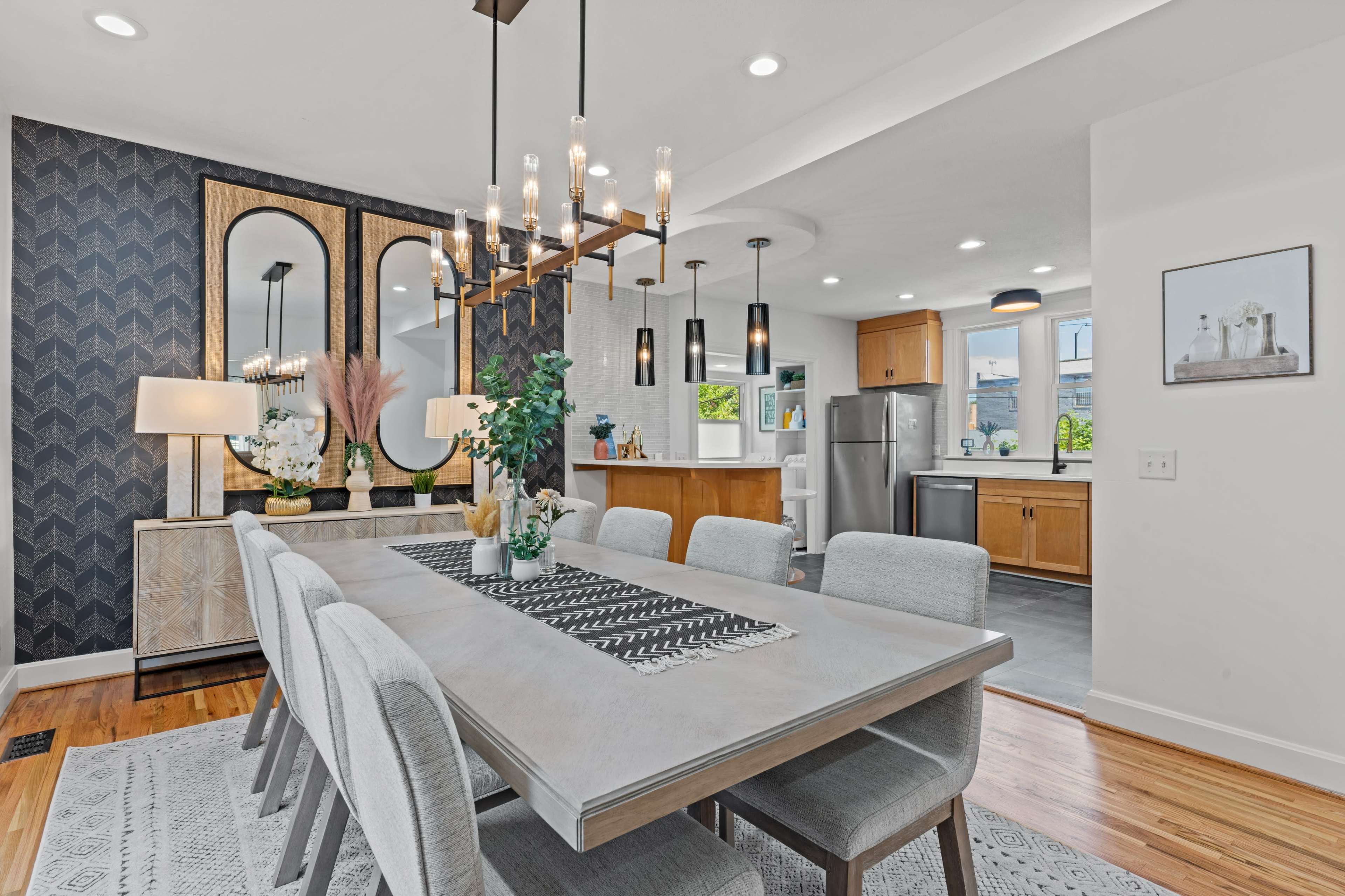 A modern dining room features a large table with six chairs, a decorative centerpiece, a stylish chandelier, and a kitchen area in the background with stainless steel appliances.