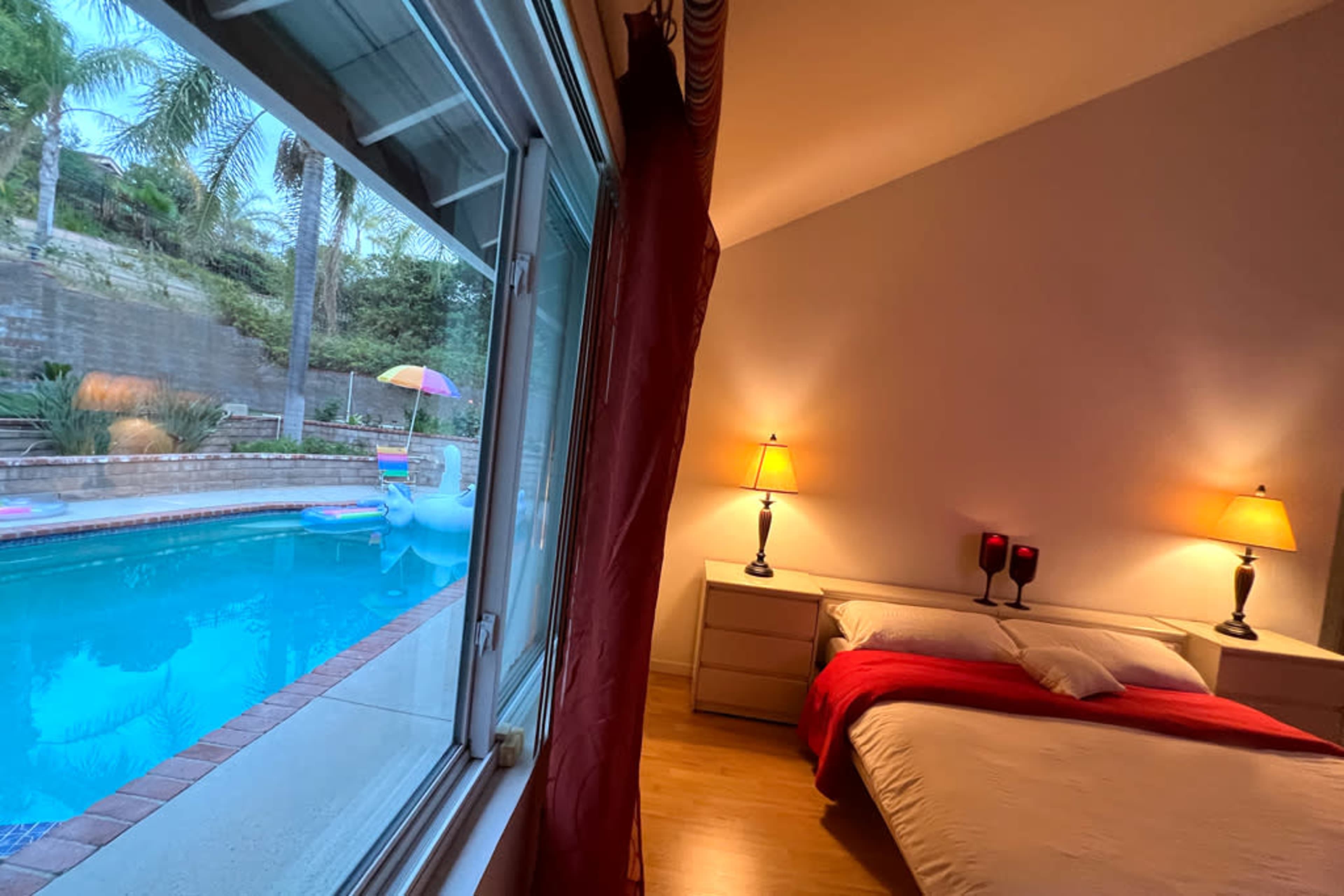 A bedroom with a bed and lamps, overlooking a pool area surrounded by palm trees.
