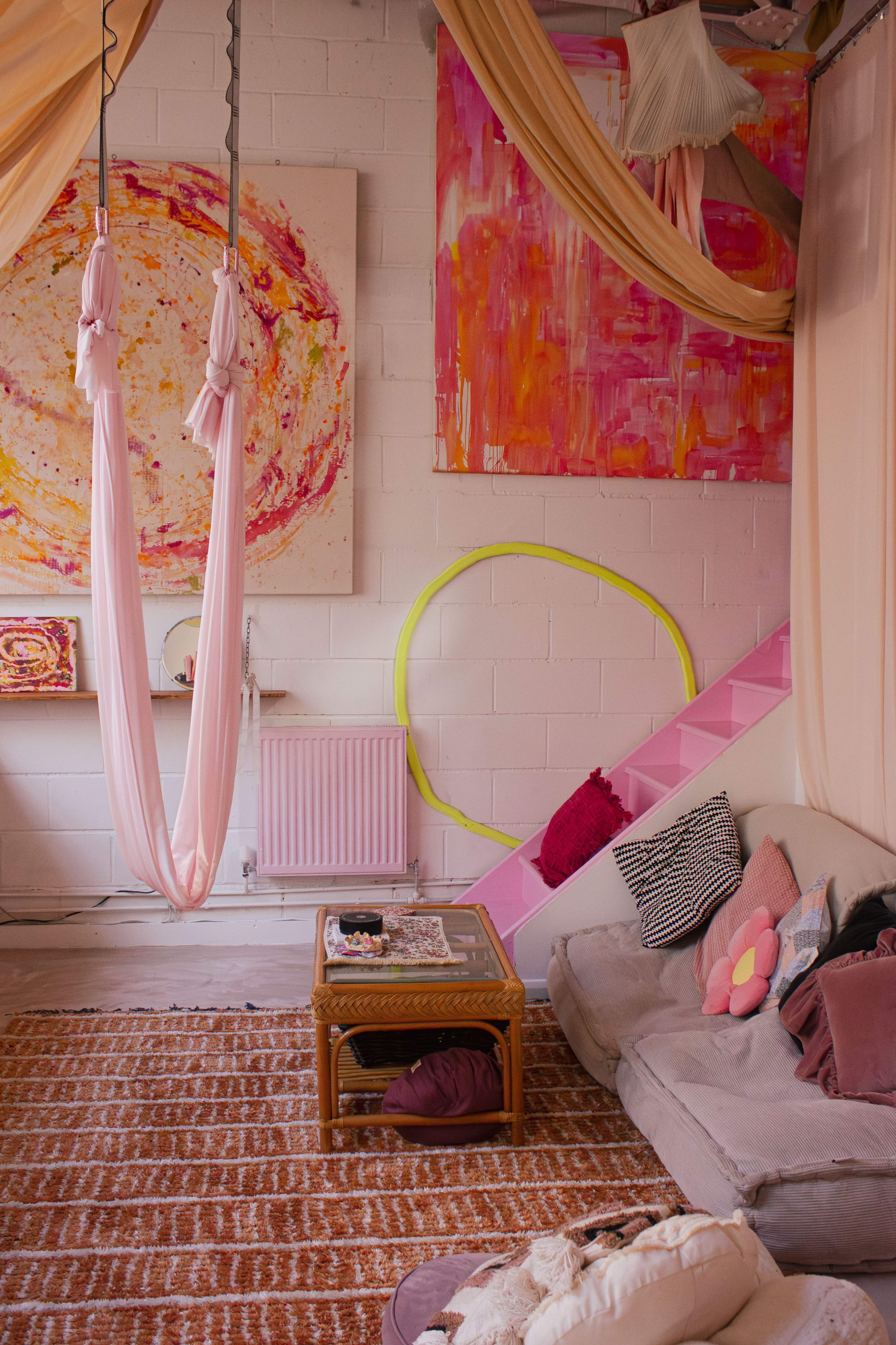 The image shows a cozy, brightly colored living space with pink fabric draped from the ceiling, a circular art piece on the wall, and a seating area featuring a light brown sofa and a small table.