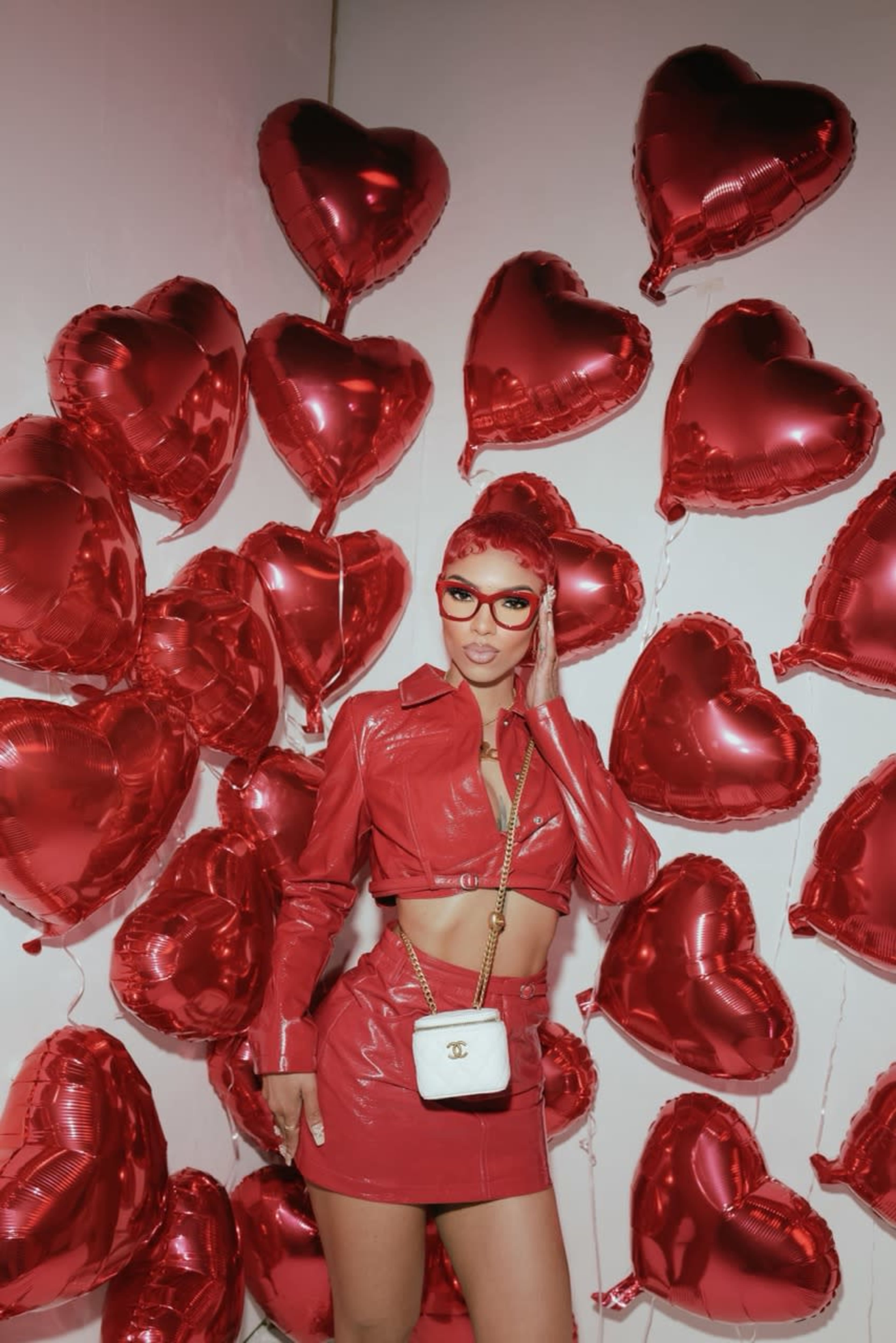 A woman in a red outfit stands in front of a wall filled with heart-shaped balloons.
