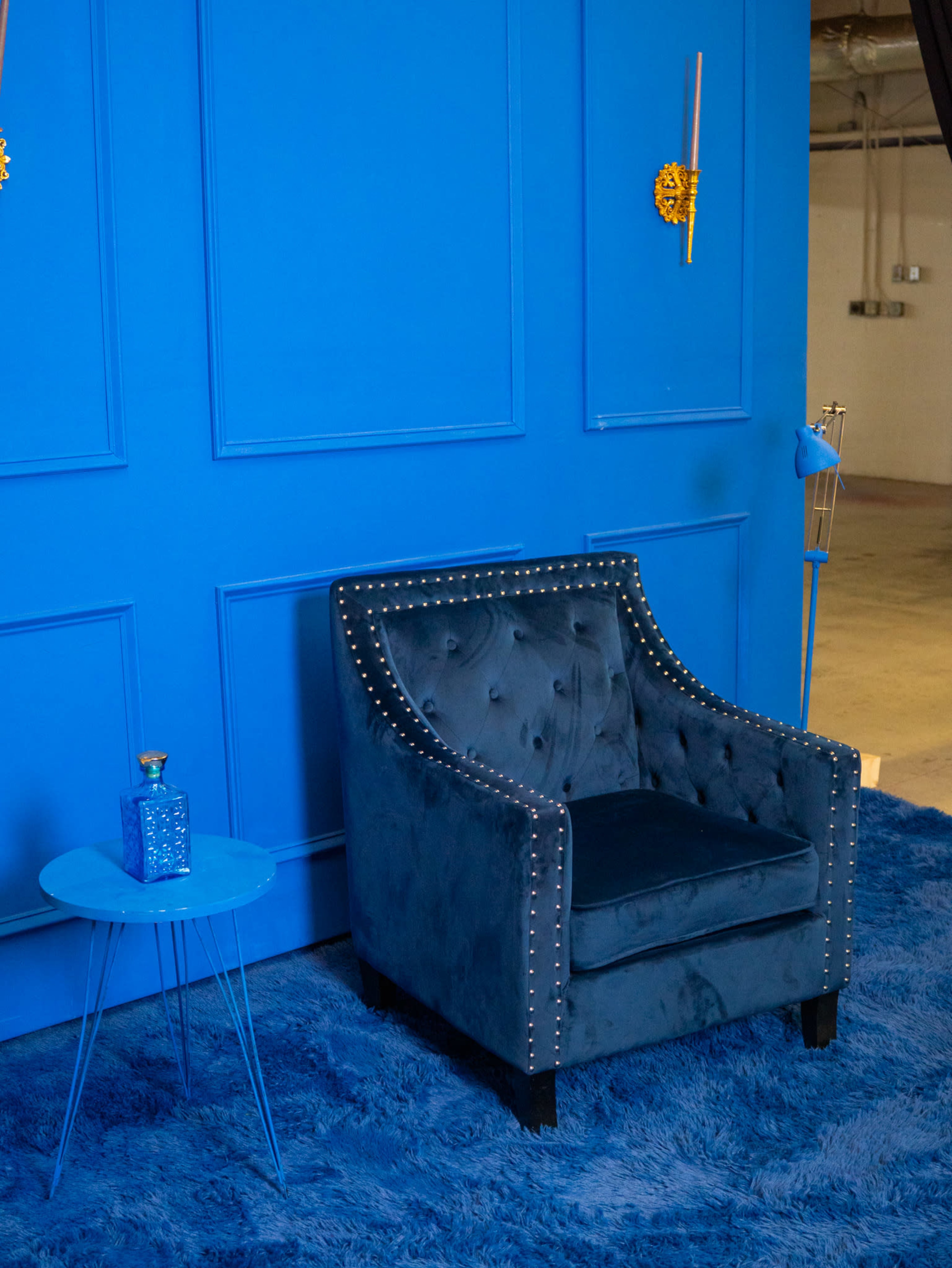 The image features a blue tufted chair with decorative studs next to a matching blue side table and set against a blue wall.