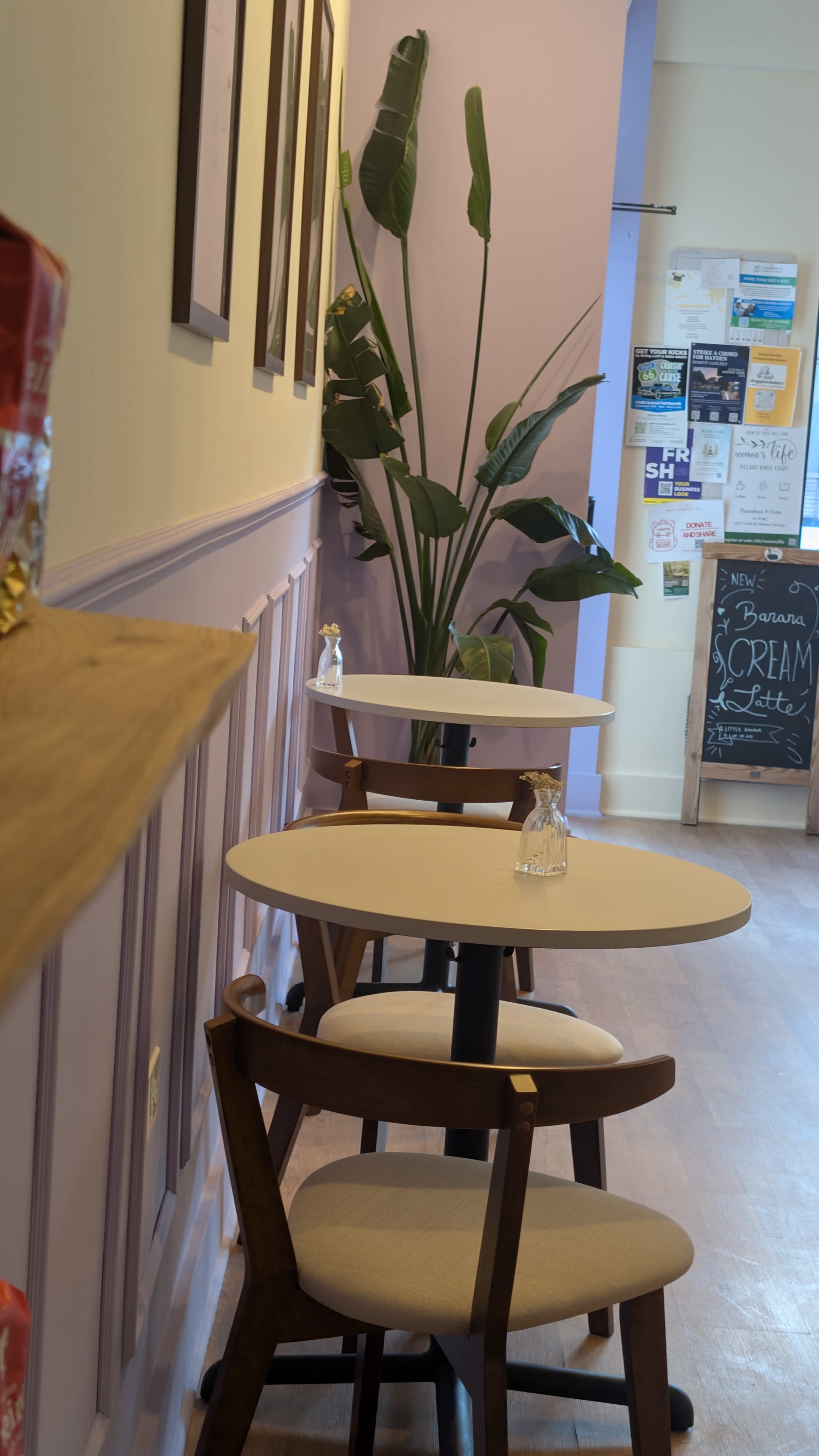 The image shows a cozy café interior with two small tables and a large plant, along with a chalkboard menu and various posters on the wall.