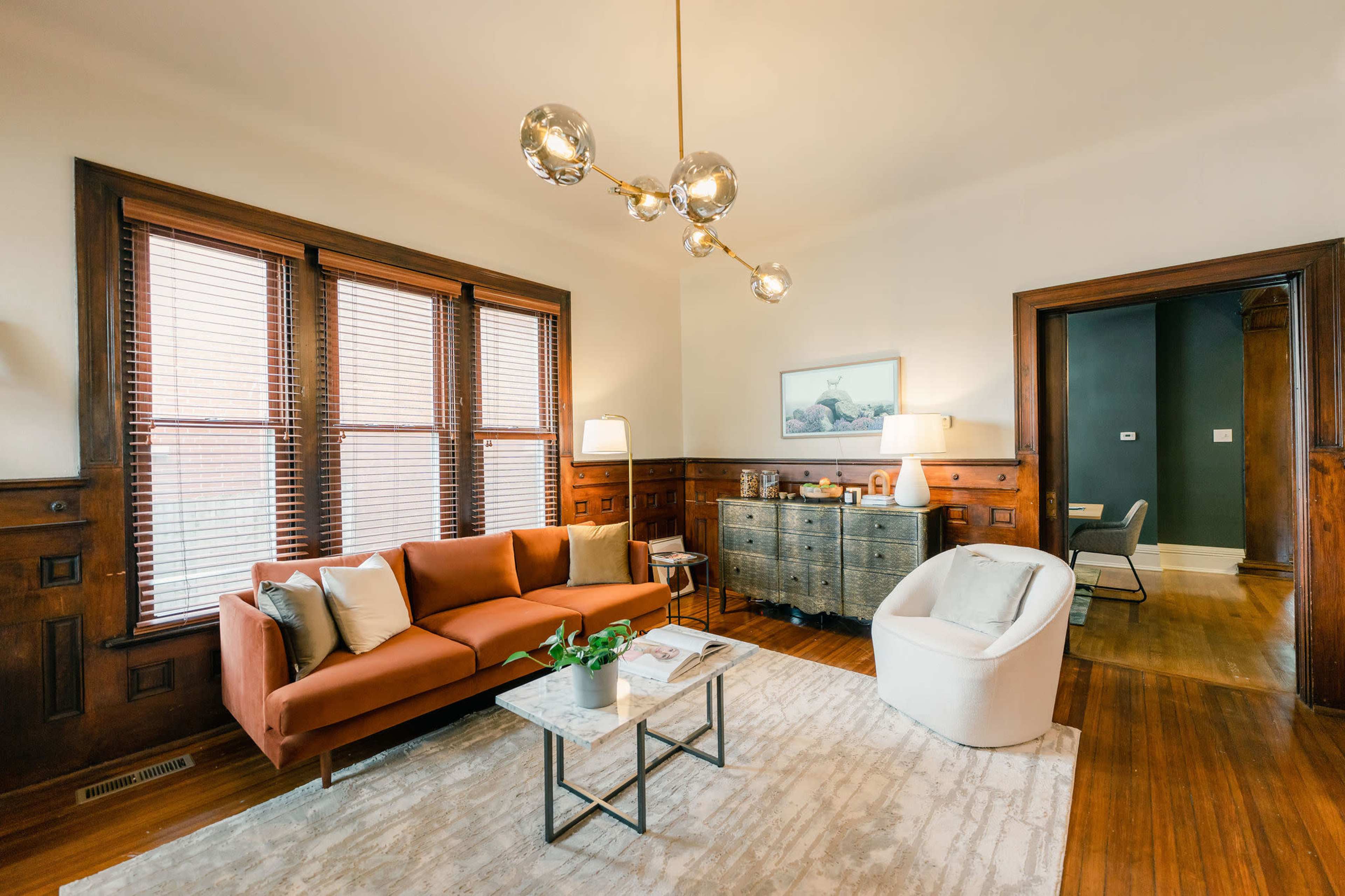 A living room features a brown sofa, a white armchair, a metal sideboard, and large windows with wooden frames.