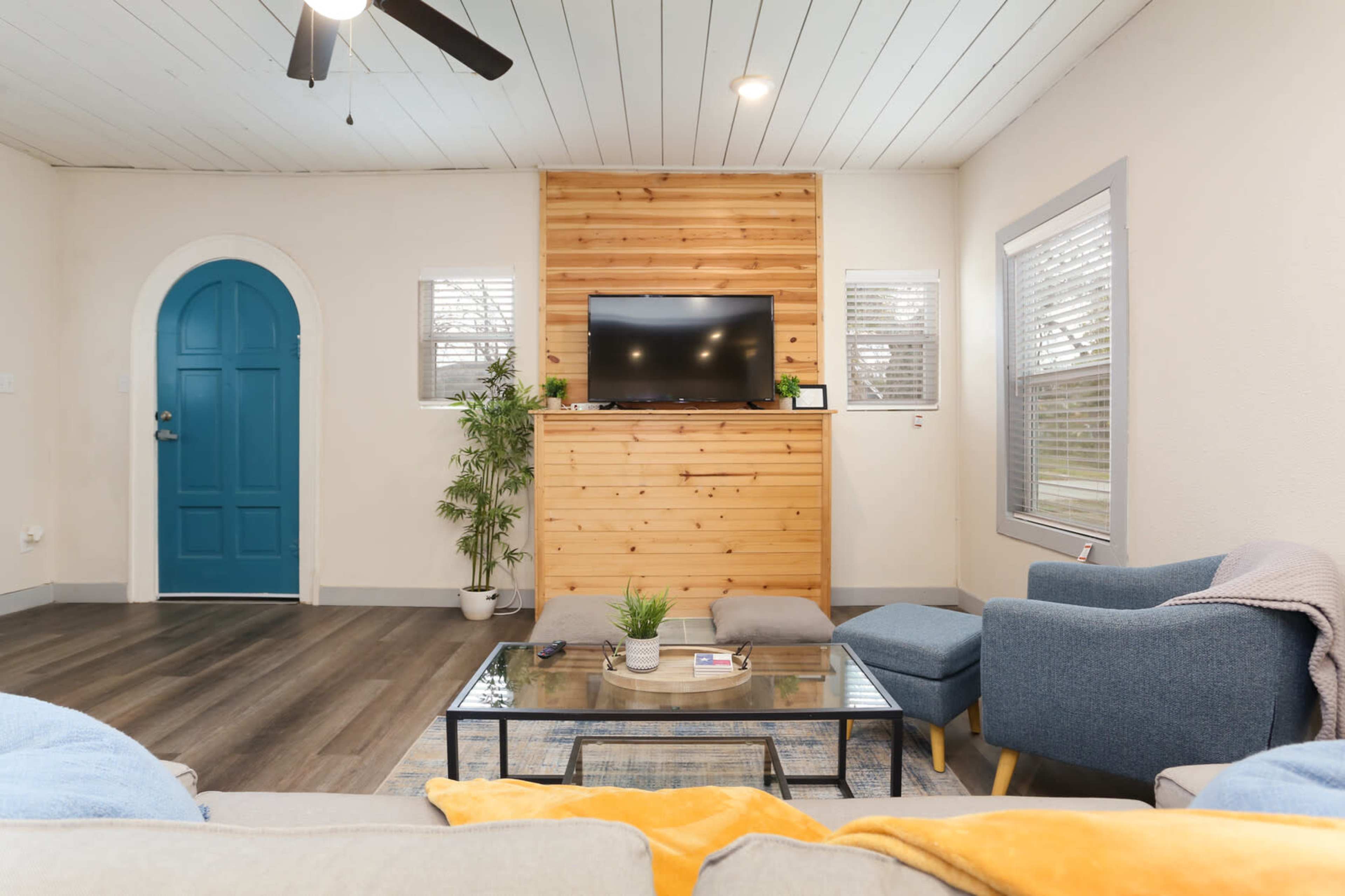 A living room features a wooden accent wall with a television, a coffee table, and blue seating, along with a door painted teal.