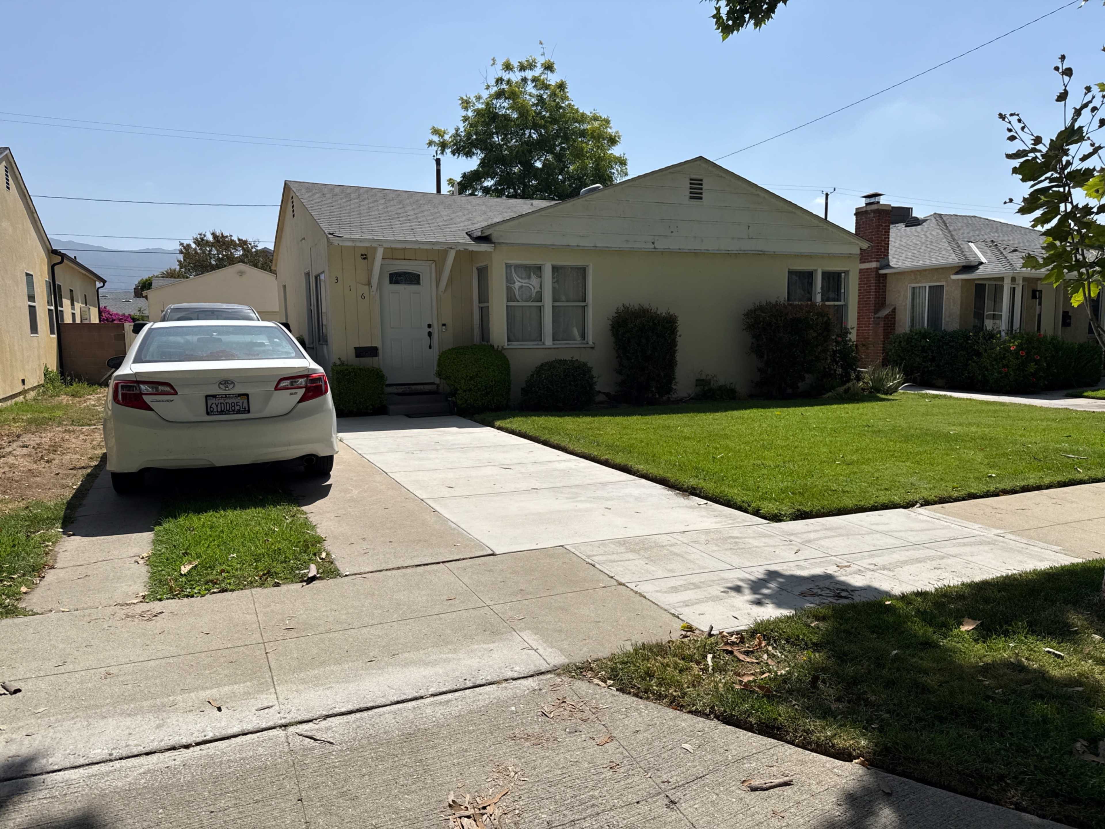 A white car is parked on the driveway of a light-colored single-story house surrounded by neatly trimmed grass and shrubs.
