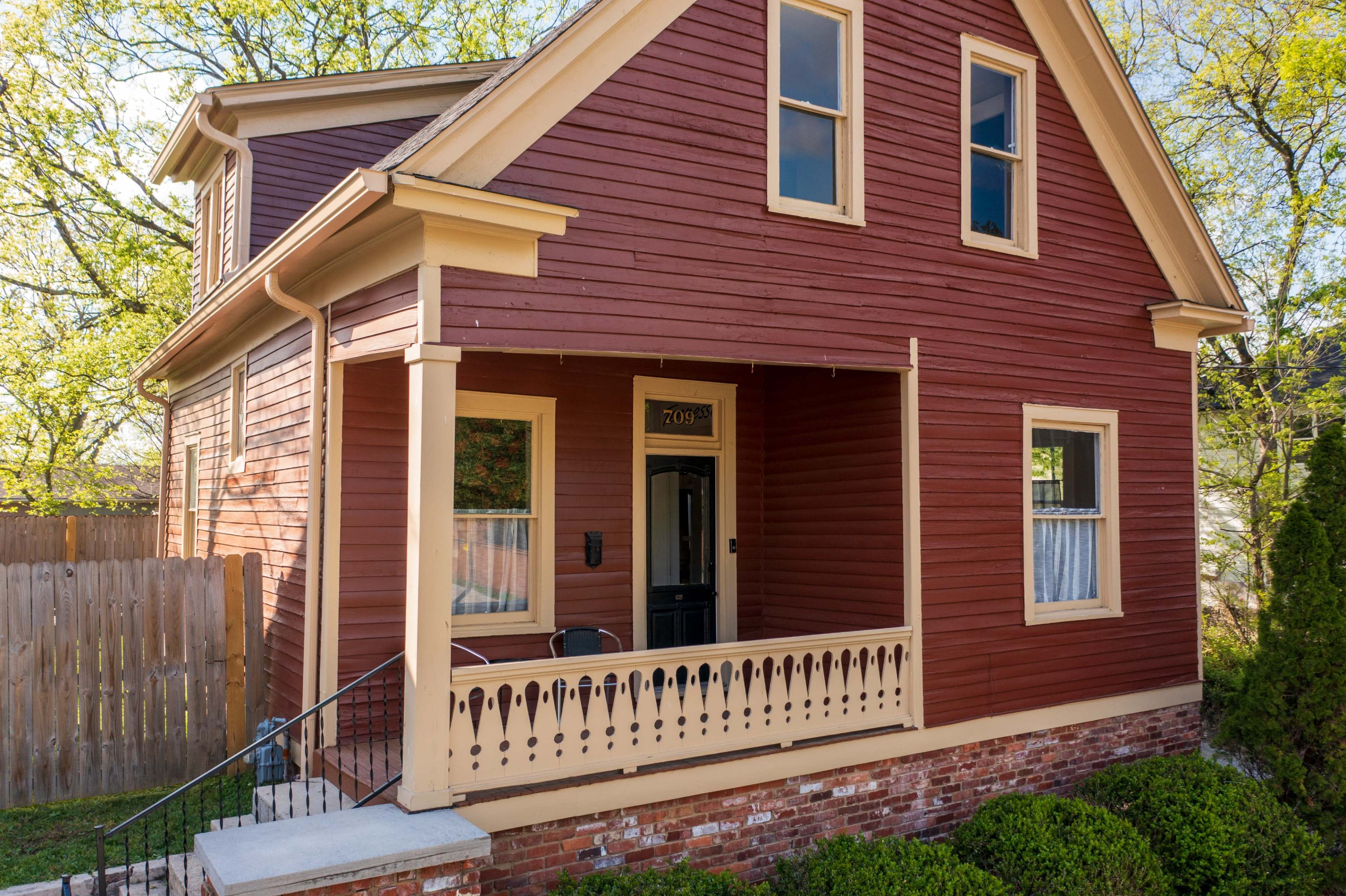 A two-story house with a red wooden exterior and a front porch features white trim and a small set of stairs leading to the entrance.