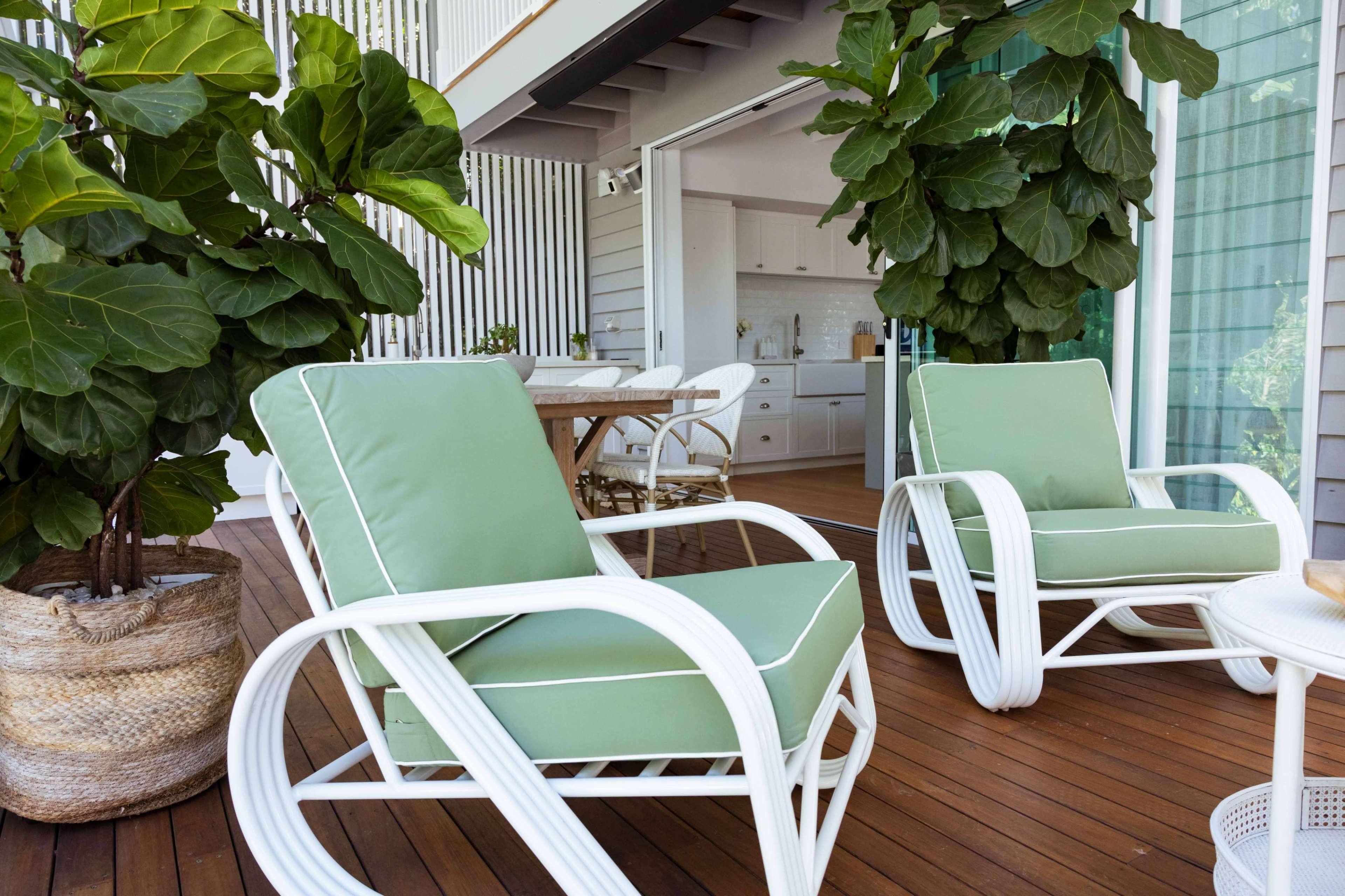 Two green cushioned chairs with white frames sit on a wooden deck, surrounded by large potted plants and a view of a modern kitchen through an open sliding door.