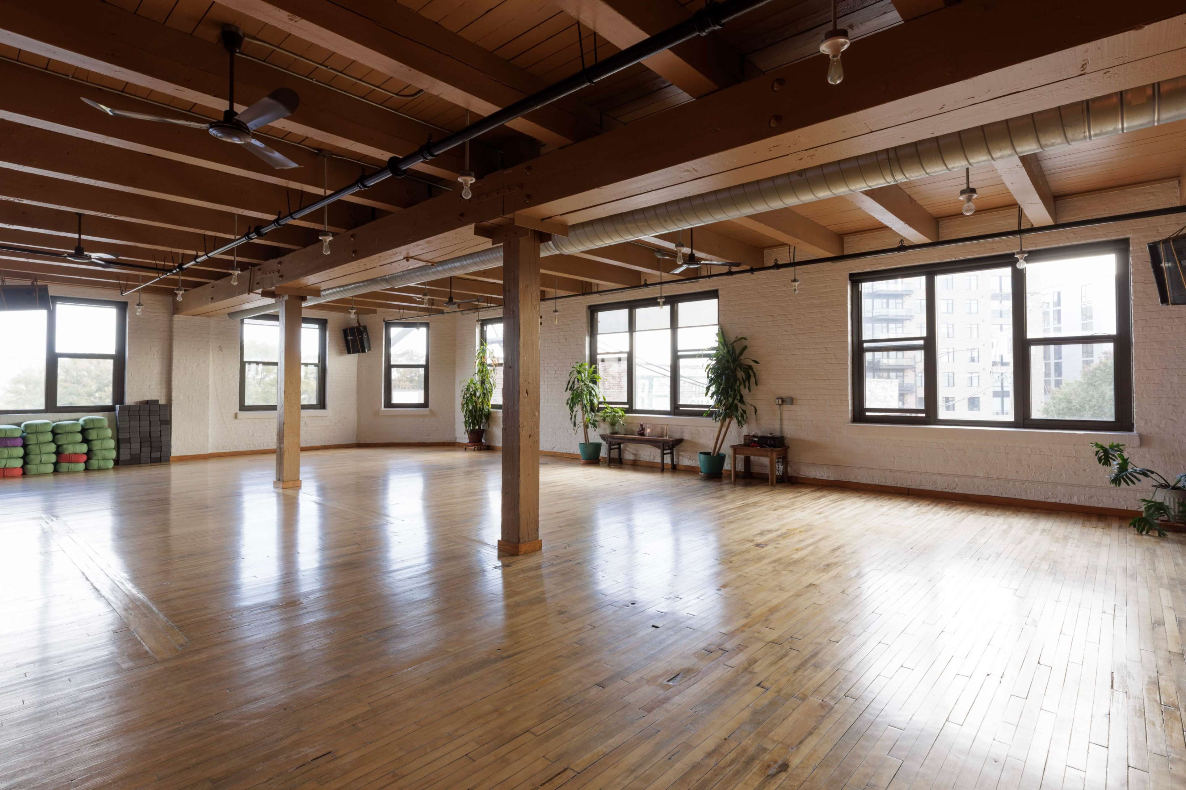 The image shows a spacious, well-lit room with wooden flooring, large windows, and potted plants, designed for activities like yoga or group classes.