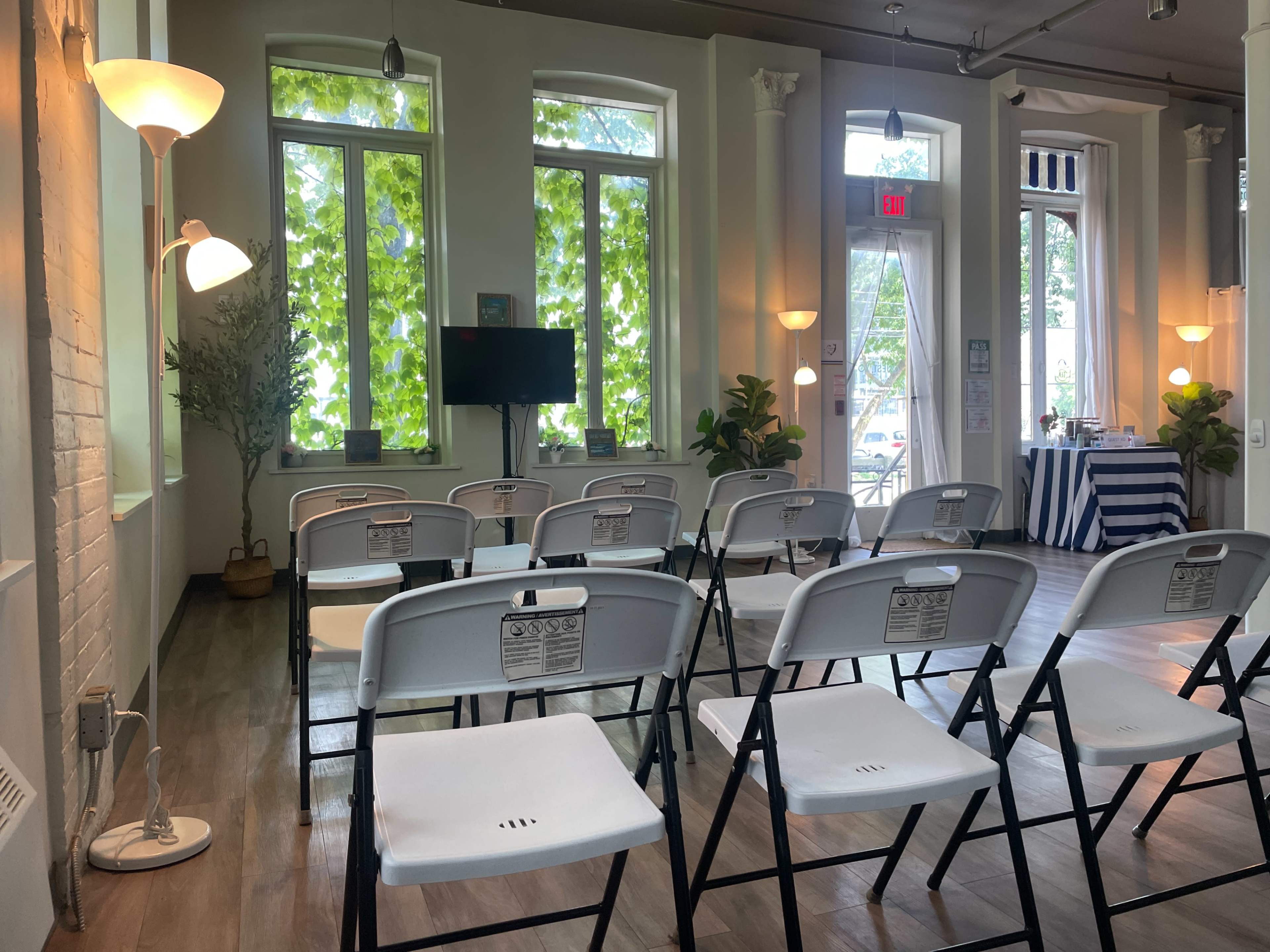 A room with folding chairs arranged in rows, large windows allowing natural light, and decorative plants providing a welcoming atmosphere.