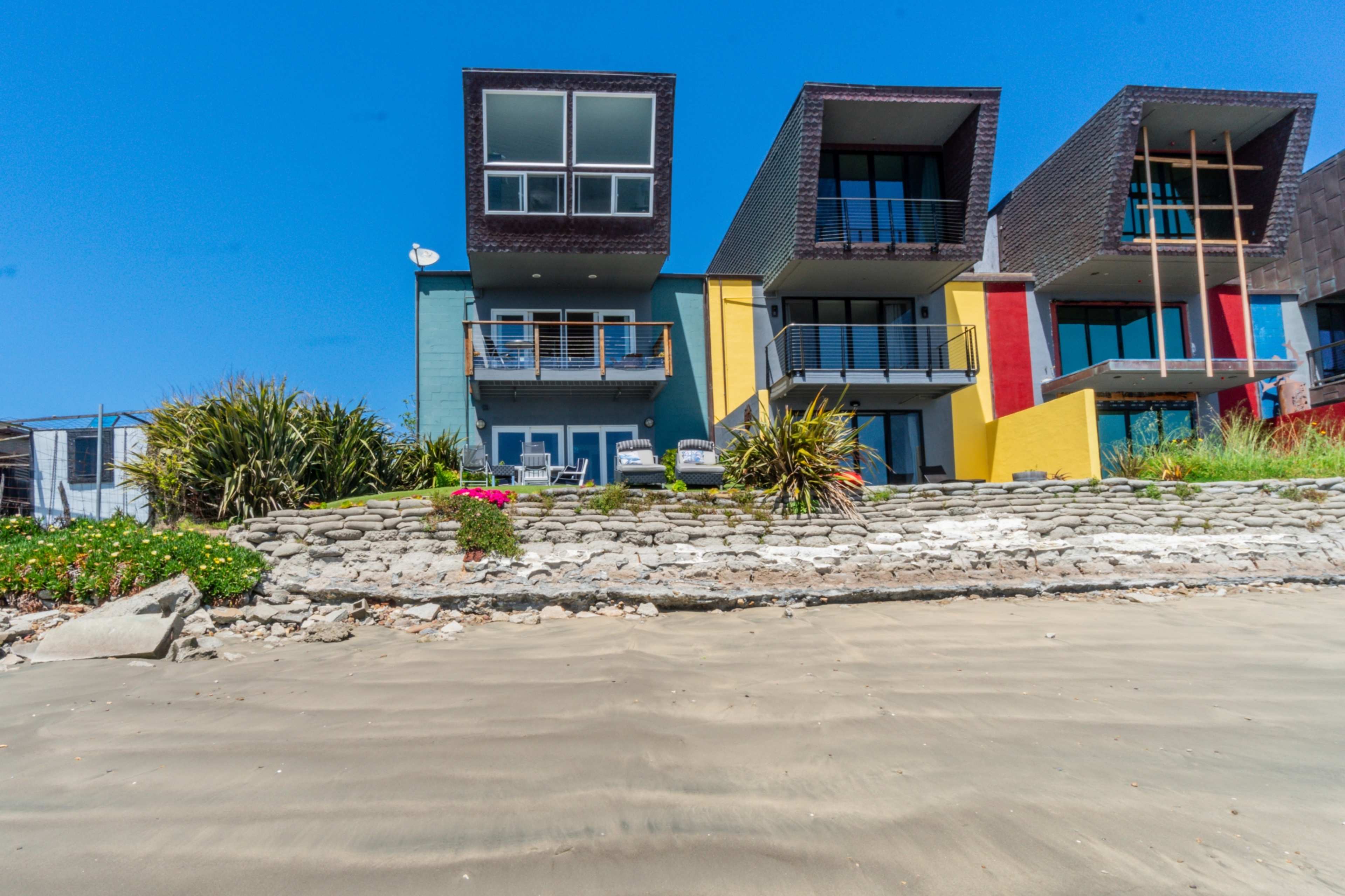 The image shows a row of modern beach houses with varying architectural designs, situated along a sandy shoreline.