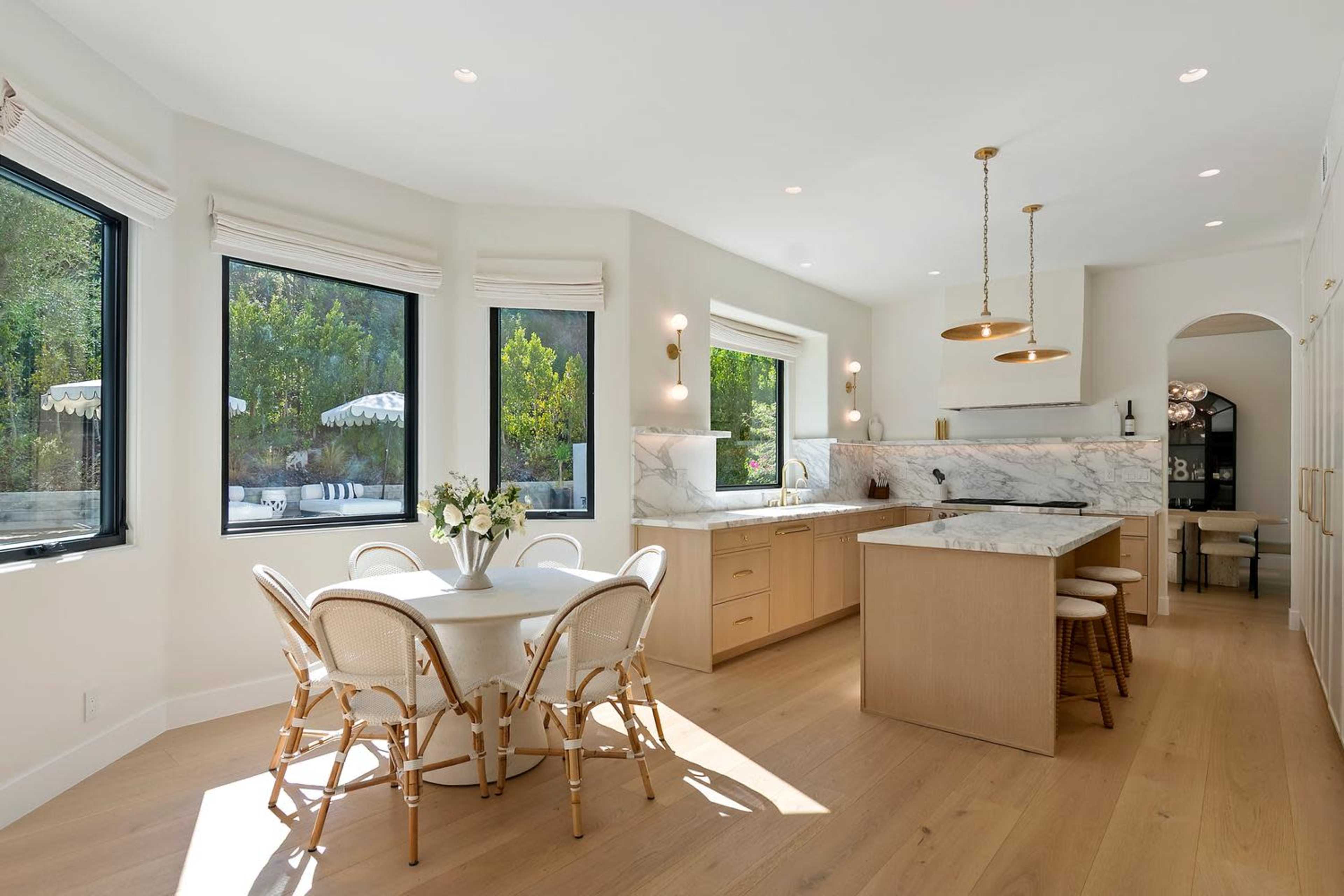 The image shows a modern kitchen featuring a large marble countertop, wooden cabinets, and a round dining table with four chairs, all set against multiple windows that allow natural light to enter.