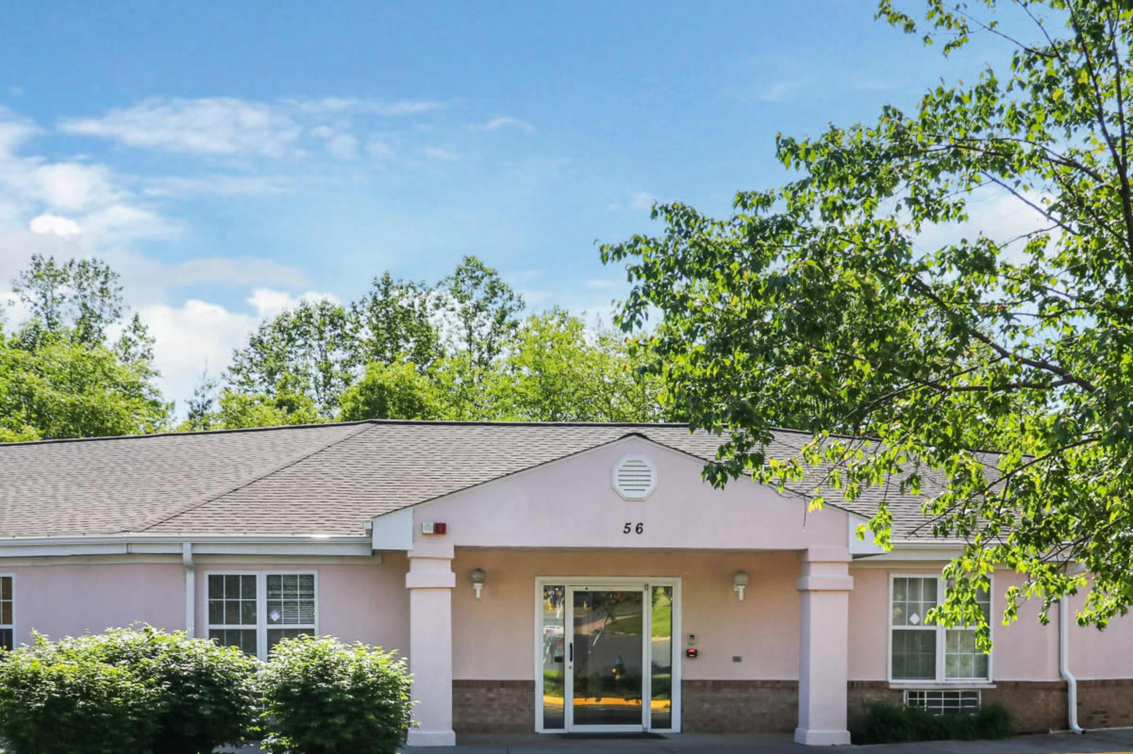 The image shows the entrance of a single-story building with a light pink exterior, framed by green trees and shrubs.