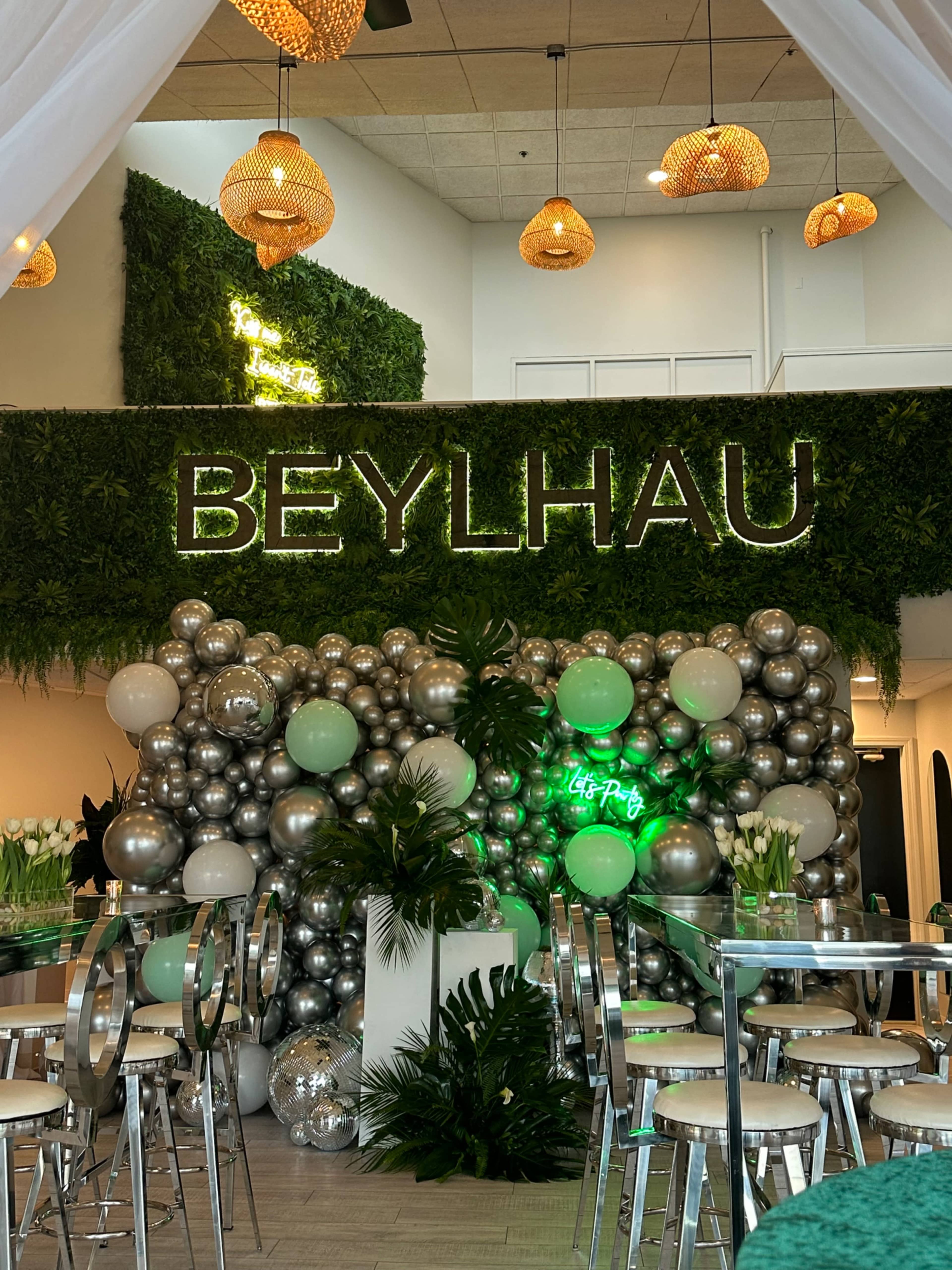 A decorated event space featuring a green wall with the word "BEYLHAU" in neon lights, surrounded by a large display of silver and green balloons.