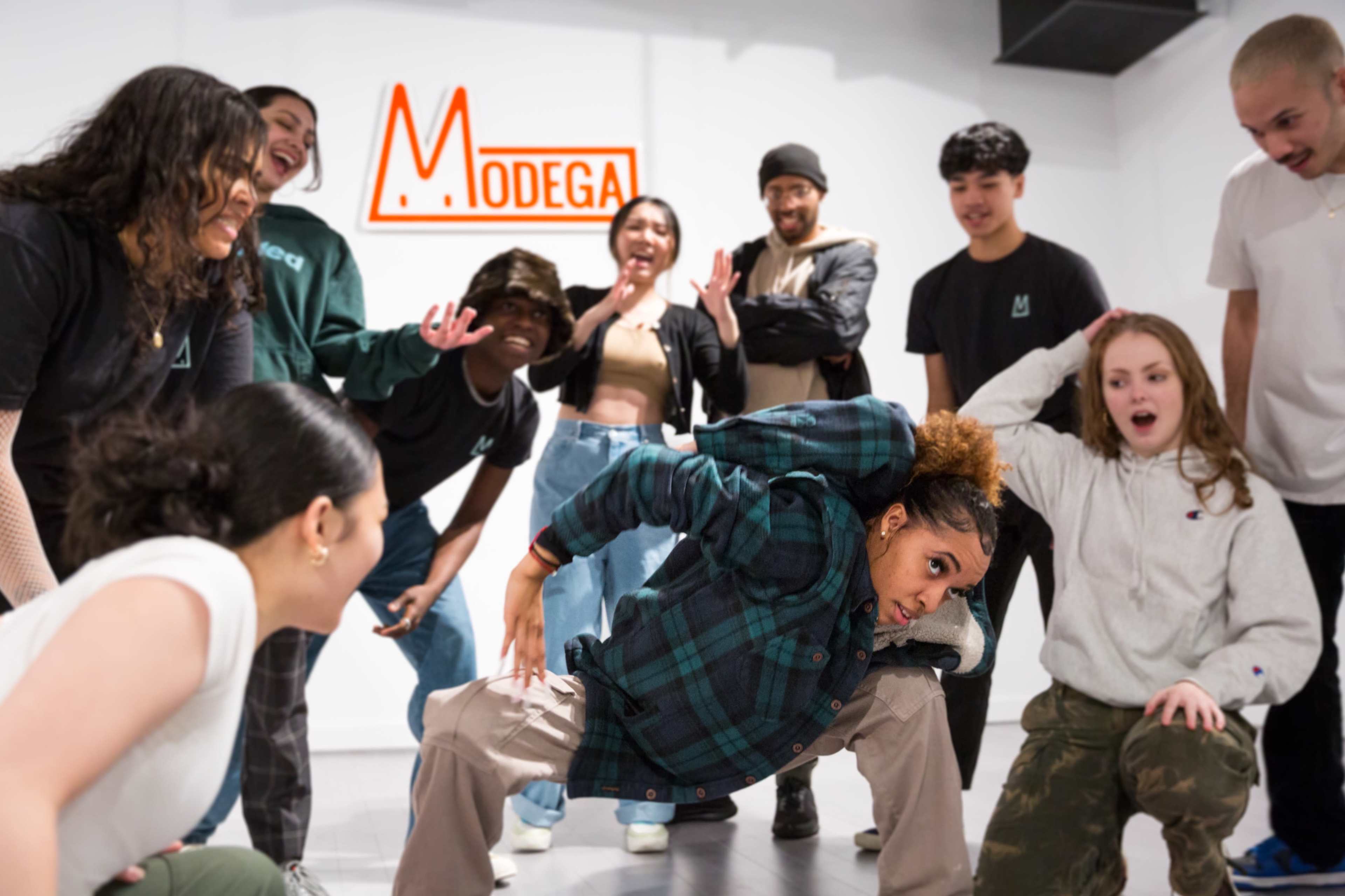 A group of dancers watches as one dancer performs a flexible pose in front of a sign that reads "Modega."