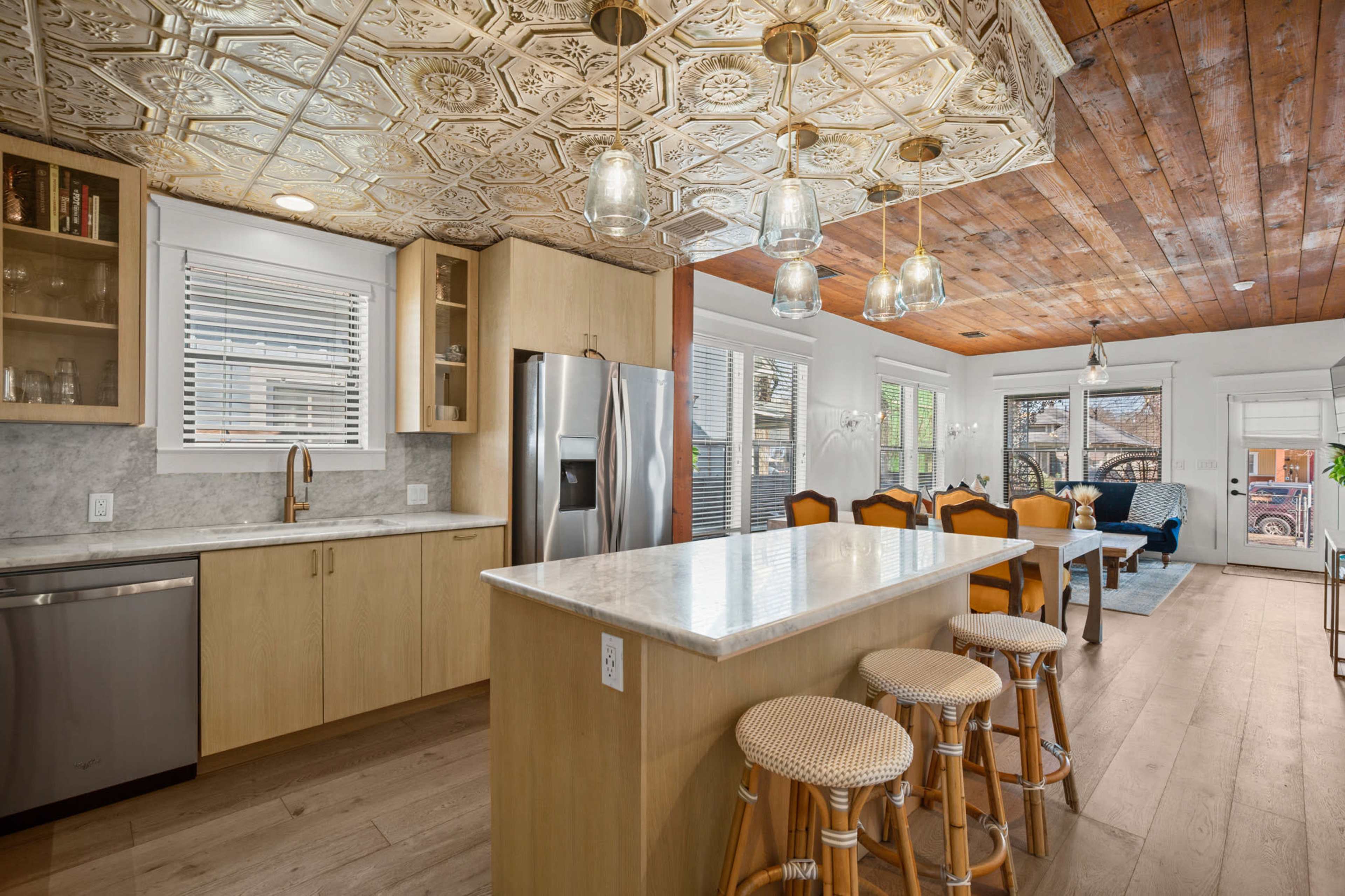 The image depicts a modern kitchen with a central island, metal light fixtures, and a dining area featuring wooden chairs and a living room visible in the background.