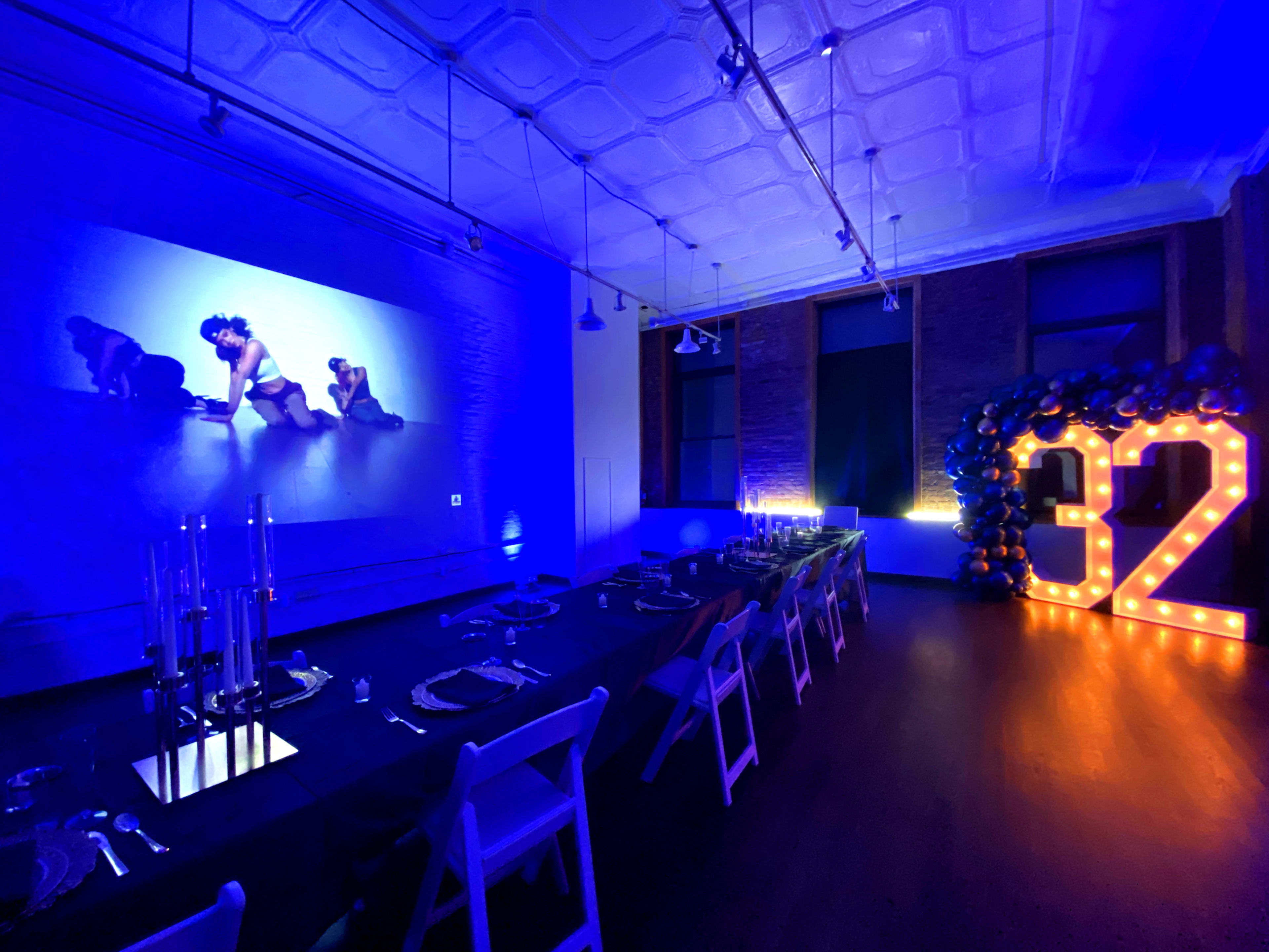 A dining area is set with a long table adorned with elegant place settings, illuminated by blue lighting and featuring a large illuminated "32" alongside a projector display.