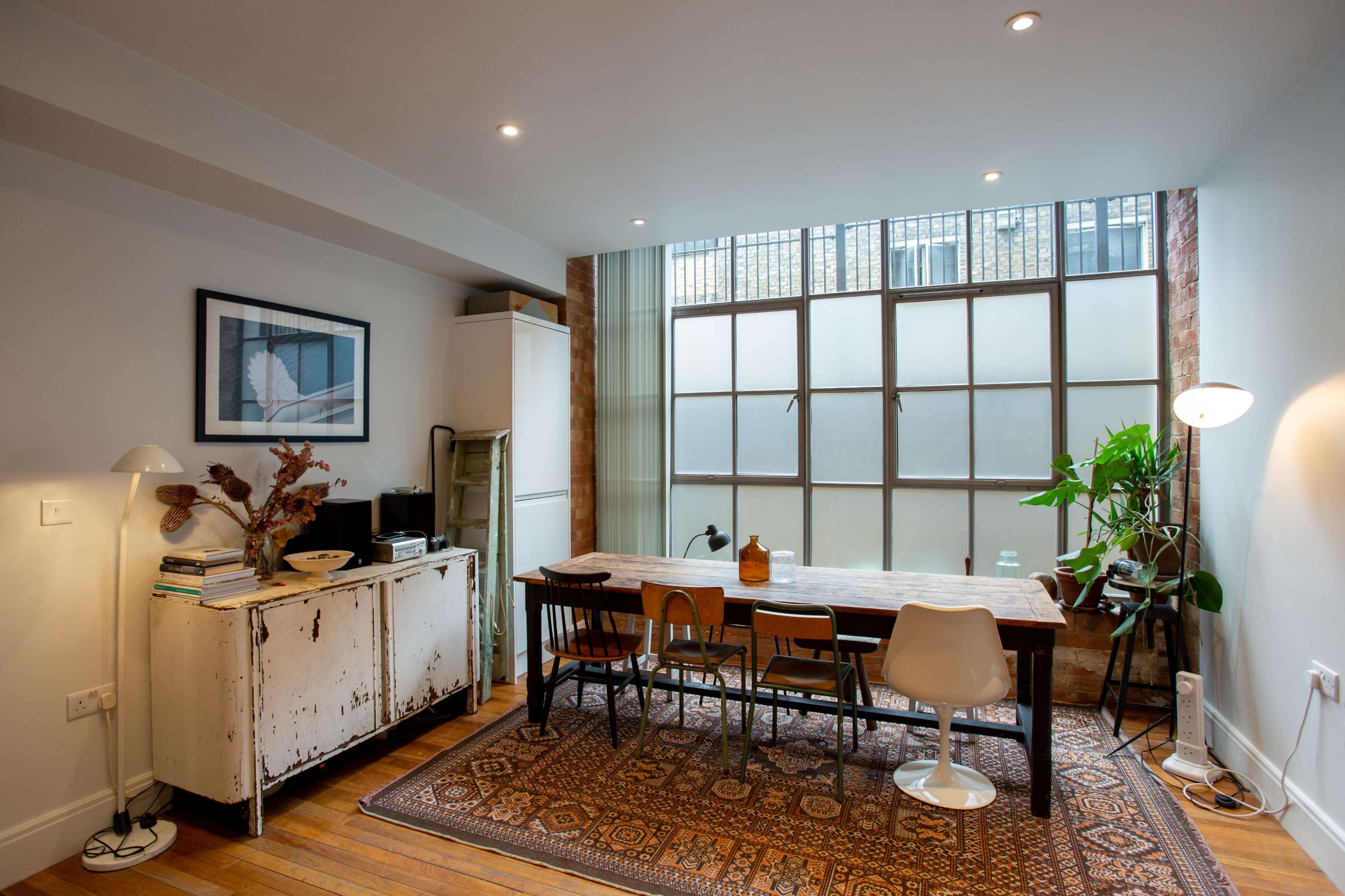 A bright dining area features a long wooden table surrounded by chairs, against a large window with a view of the outdoors.