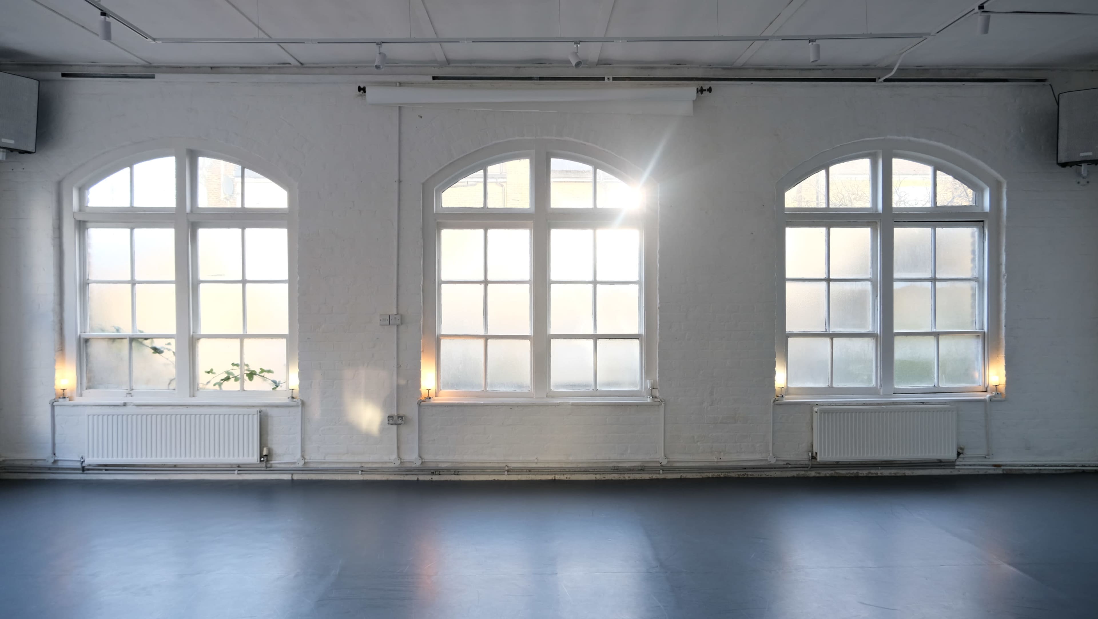 The image shows a bright, airy room with three arched windows that have soft natural light coming through and a minimalist design featuring white walls and a dark floor.