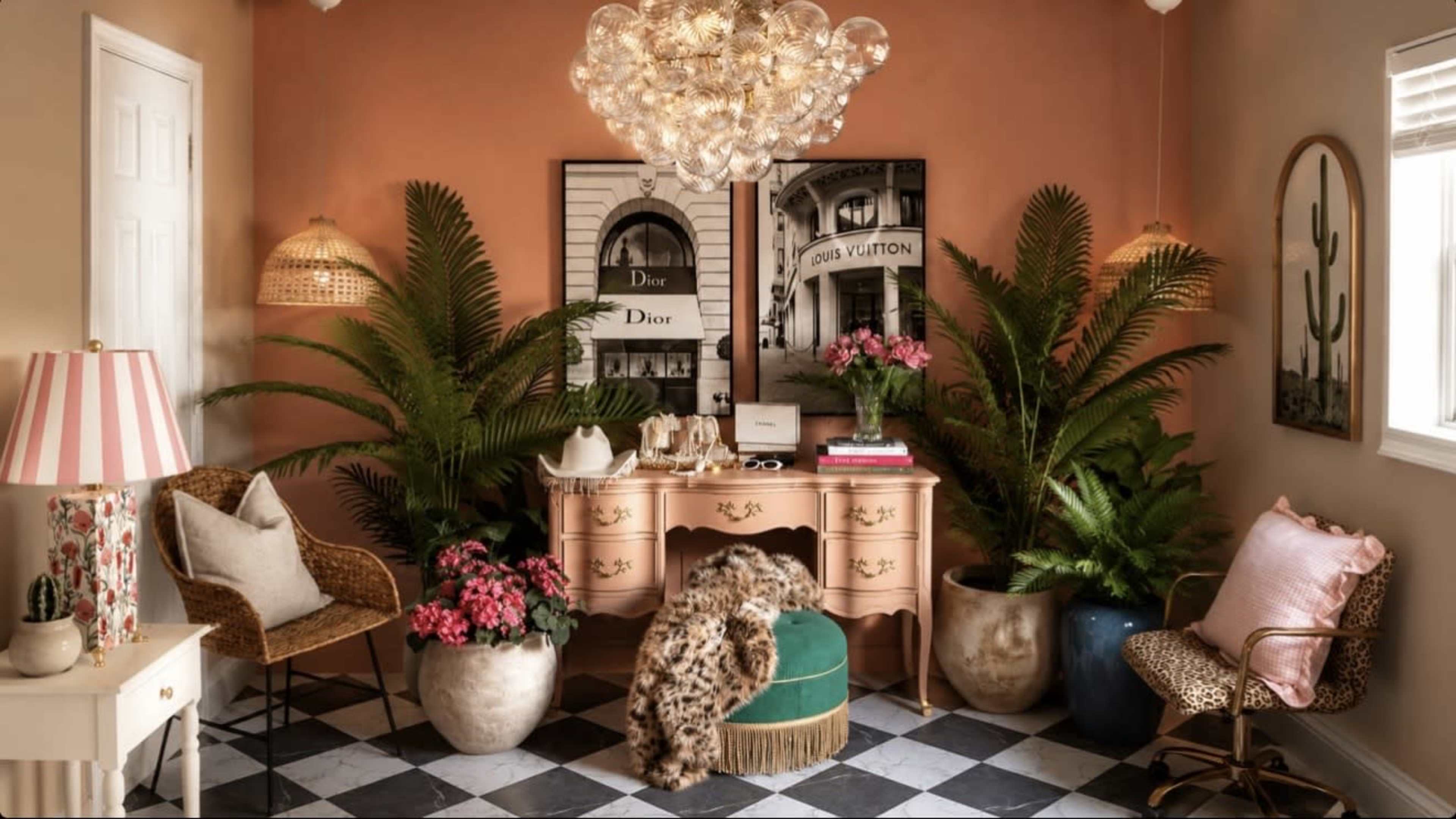 The image shows a stylish corner of a room featuring a pink and gold desk, decorative plants, and various pieces of furniture arranged harmoniously against a soft orange wall.