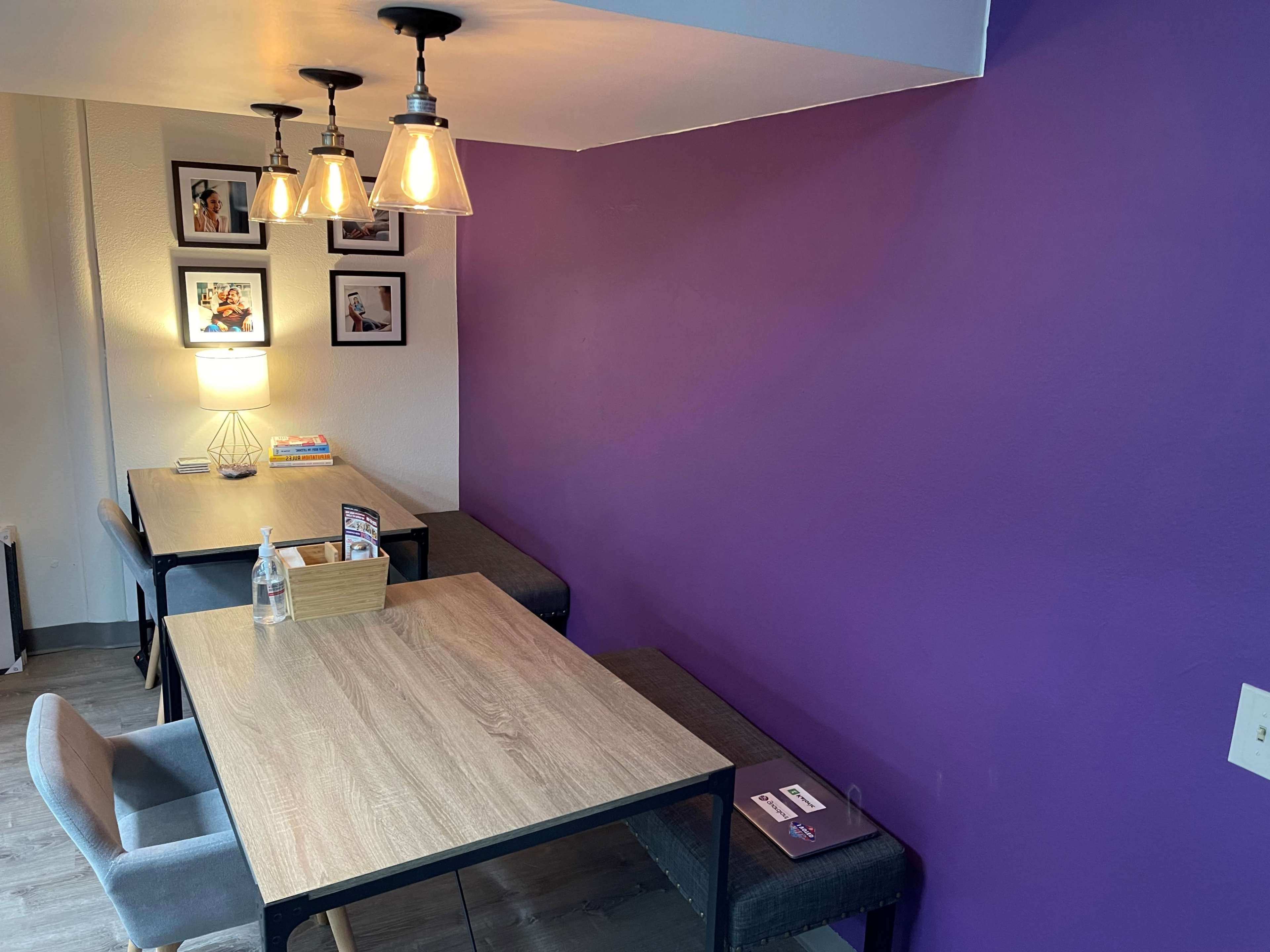 A cozy seating area with two tables, each equipped with minimalist chairs, illuminated by pendant lights, and featuring a vibrant purple wall adorned with framed pictures.