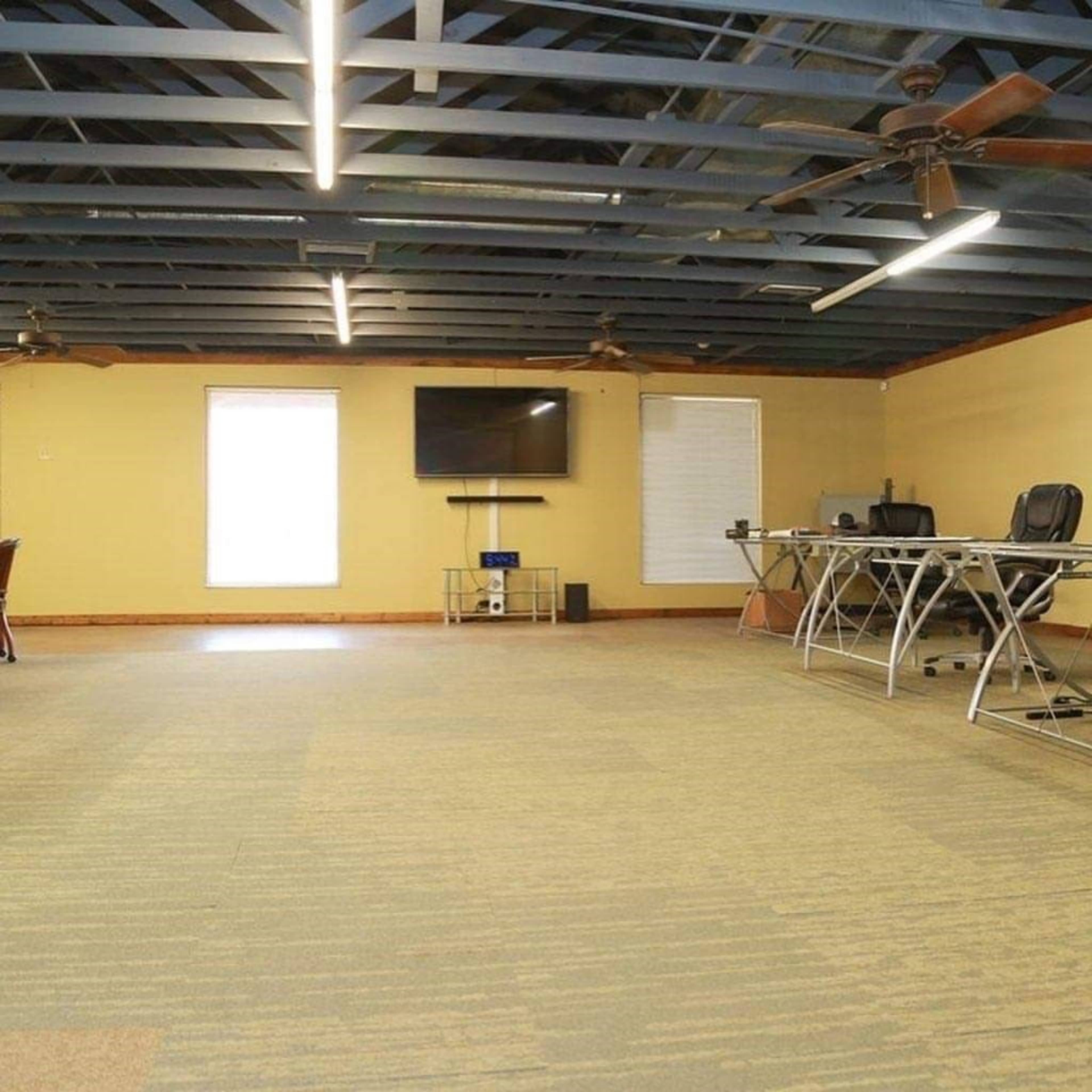 The room features a yellow wall, blue ceiling beams, a television mounted on one wall, and two workstations with chairs set up in a spacious layout.