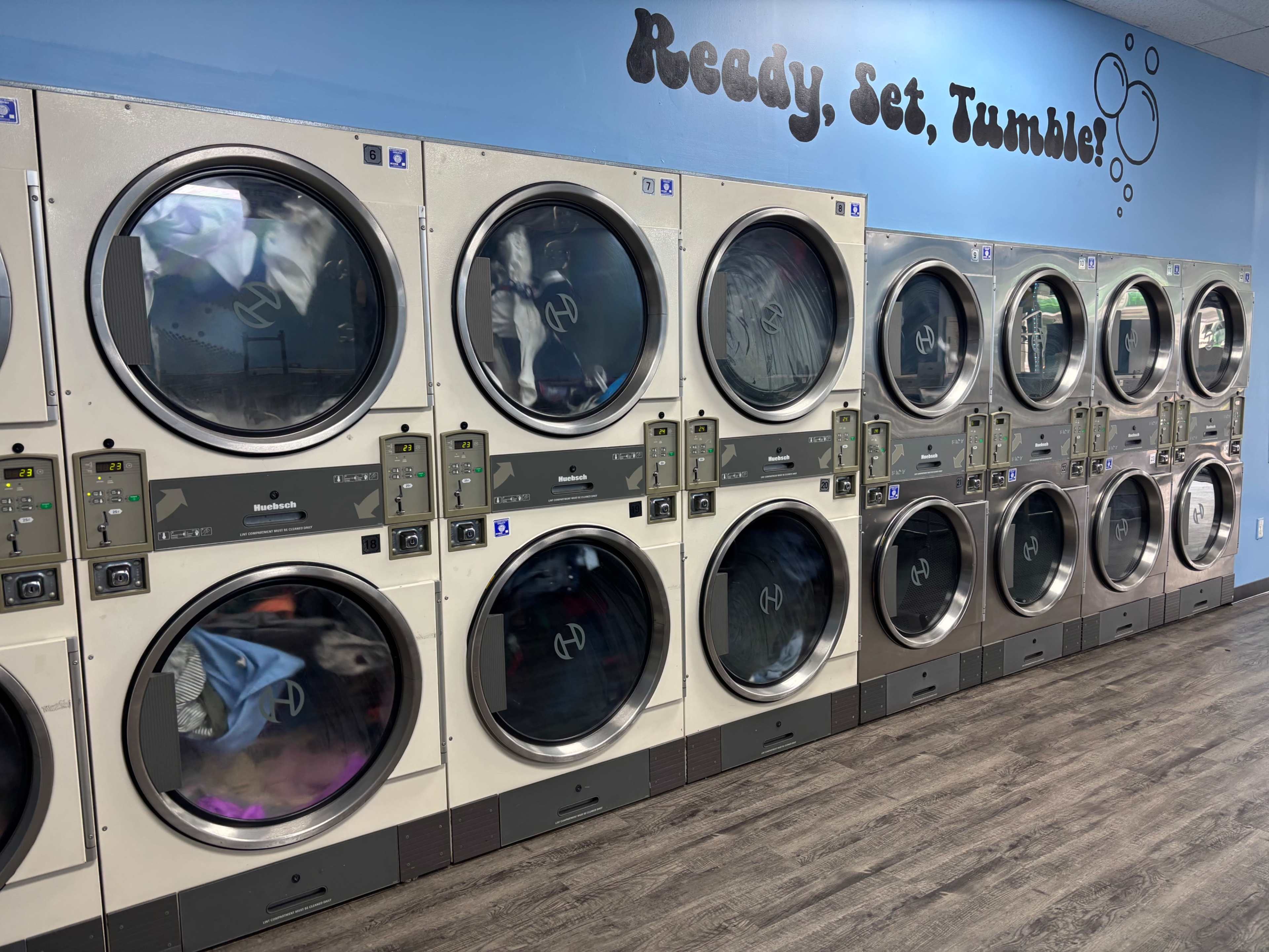A row of stacked commercial dryers is lined up against a blue wall in a laundromat.