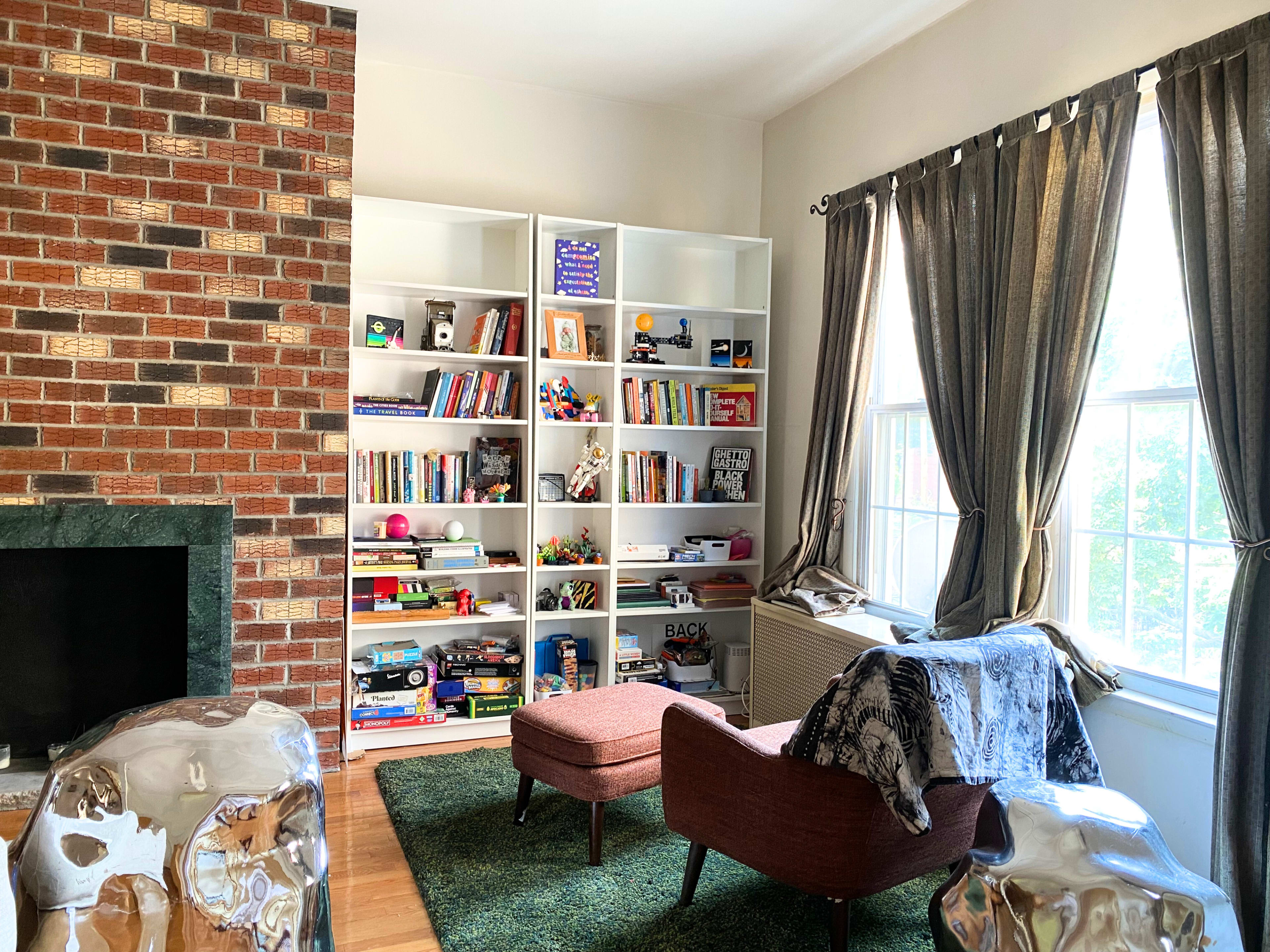 A cozy corner of a room features a brick fireplace, a bookshelf filled with colorful books and toys, and a pair of armchairs on a green rug near a window with long curtains.