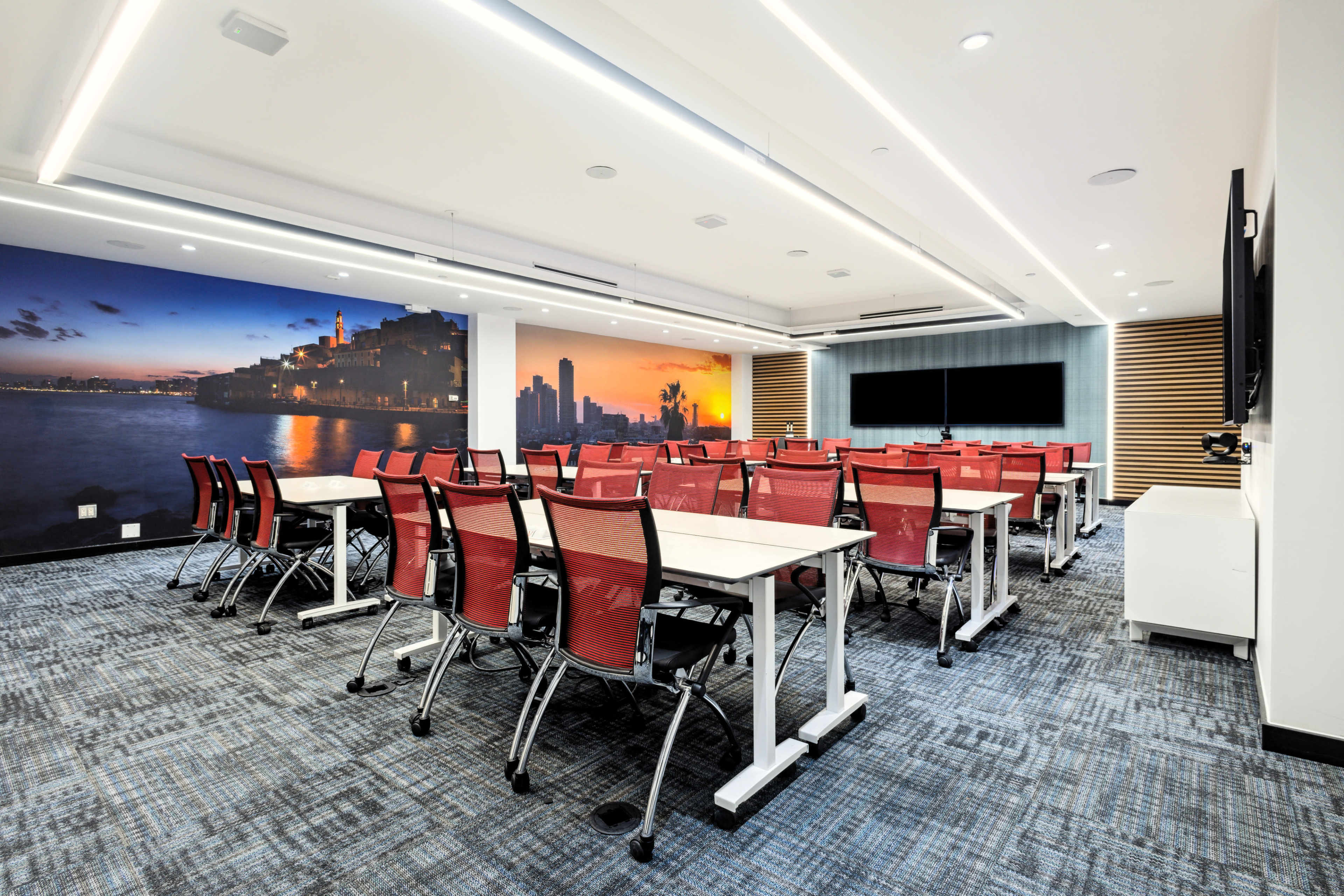 The image shows a modern conference room with multiple red and white tables and chairs arranged in a classroom style, featuring a large screen and a wall mural depicting a city skyline at sunset.