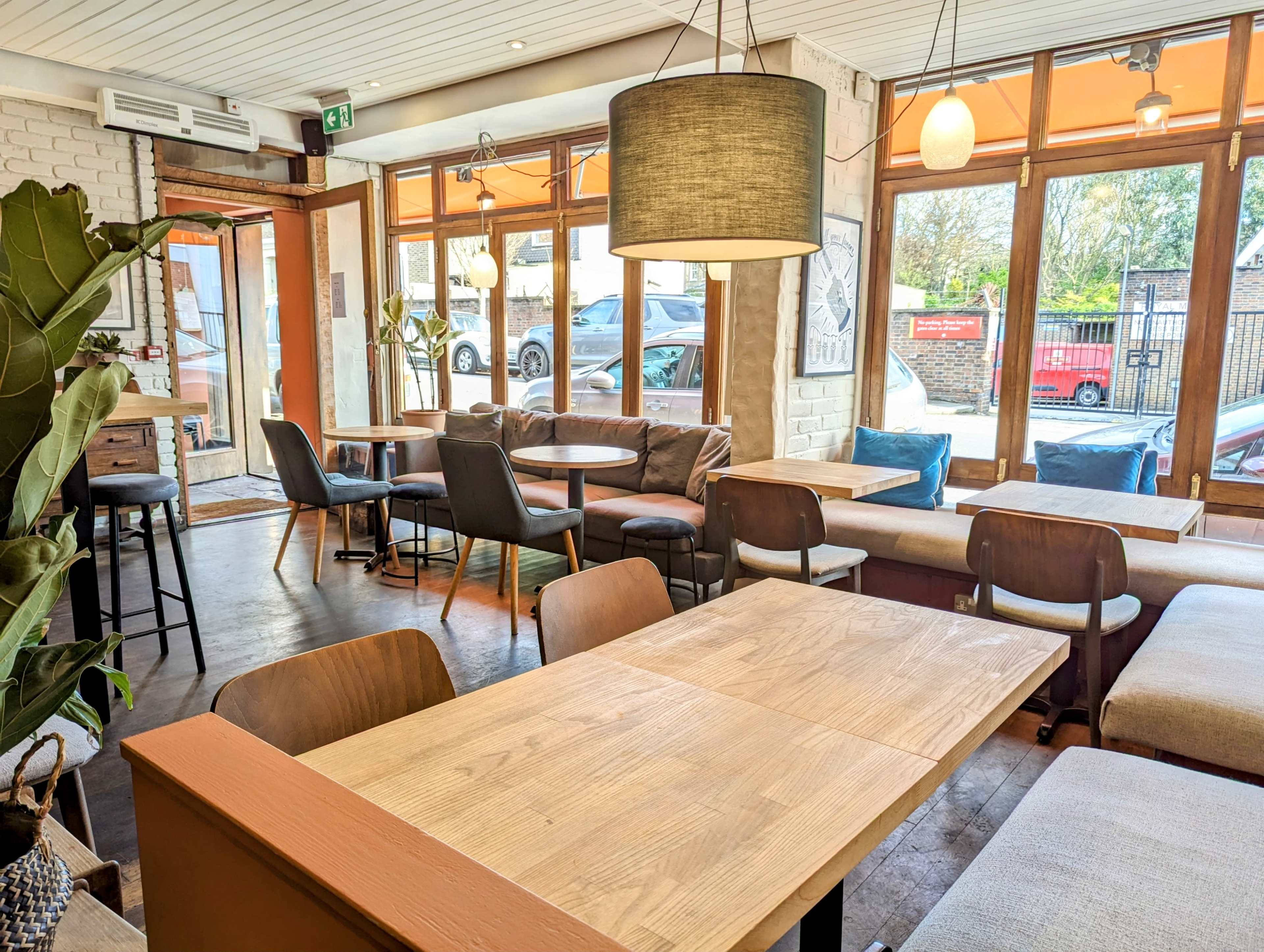 The image shows a cozy café interior with wooden tables, various seating options, and large windows allowing natural light to enter.