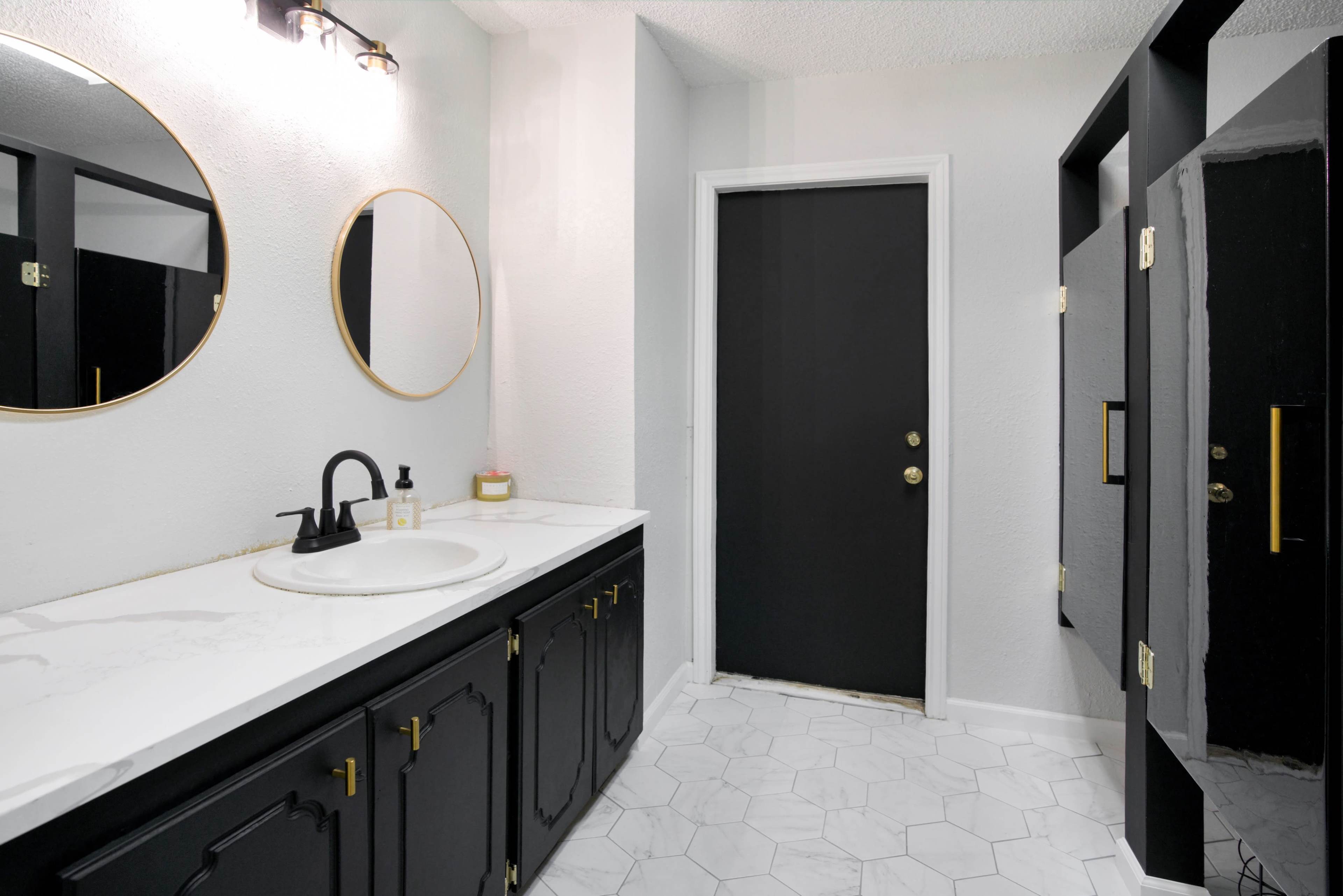 A bathroom features a black cabinetry with a white countertop, round mirrors, and a closed door leading outside, complemented by hexagonal tiled flooring.