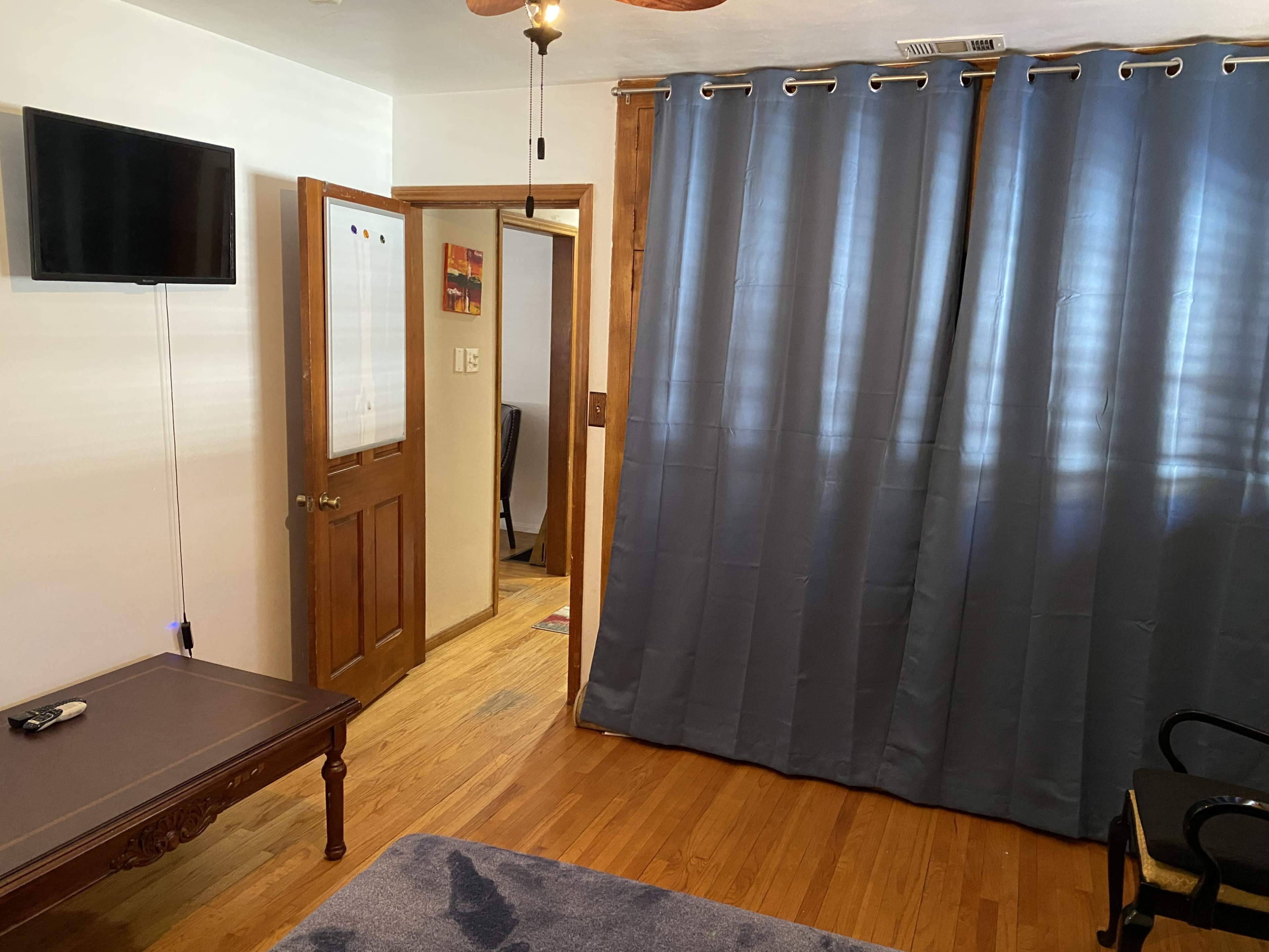 The image shows a room with wooden floors, a television mounted on the wall, a dark curtain covering a doorway, and a small table with a remote control.