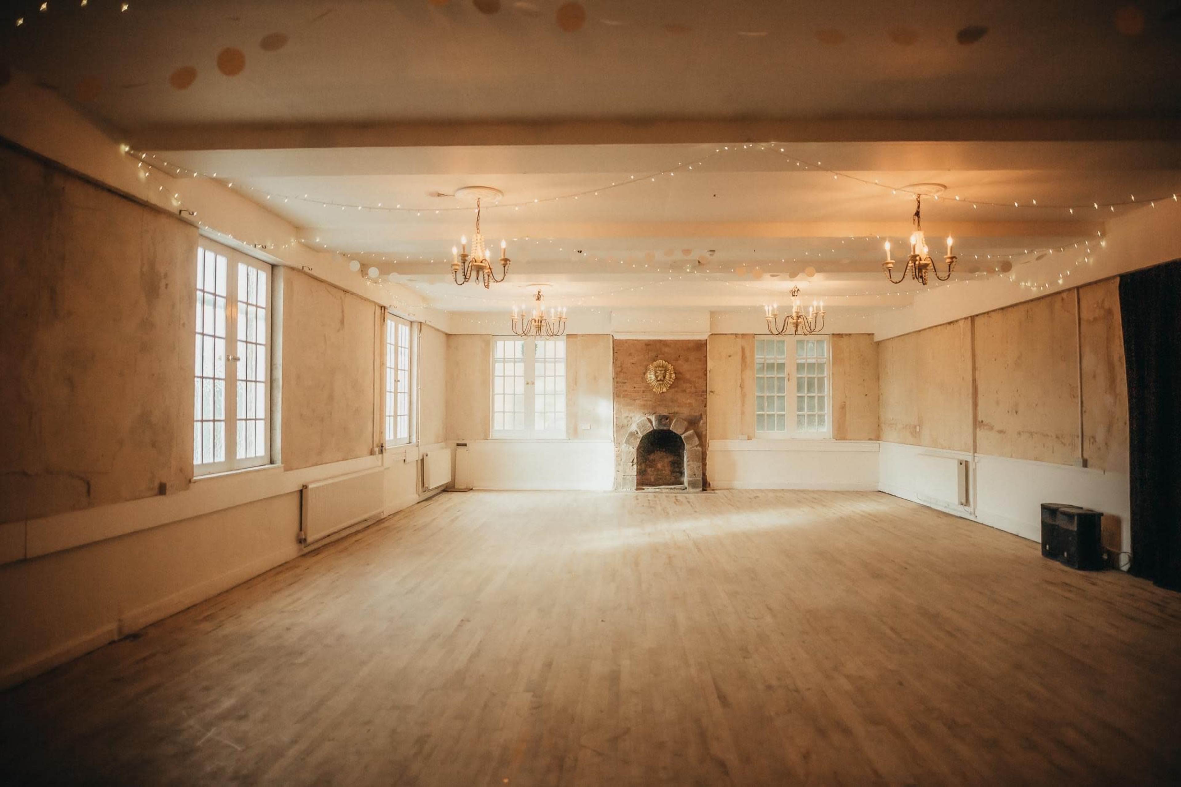 A spacious, empty room with wooden flooring, featuring large windows, a fireplace, and decorative chandeliers.