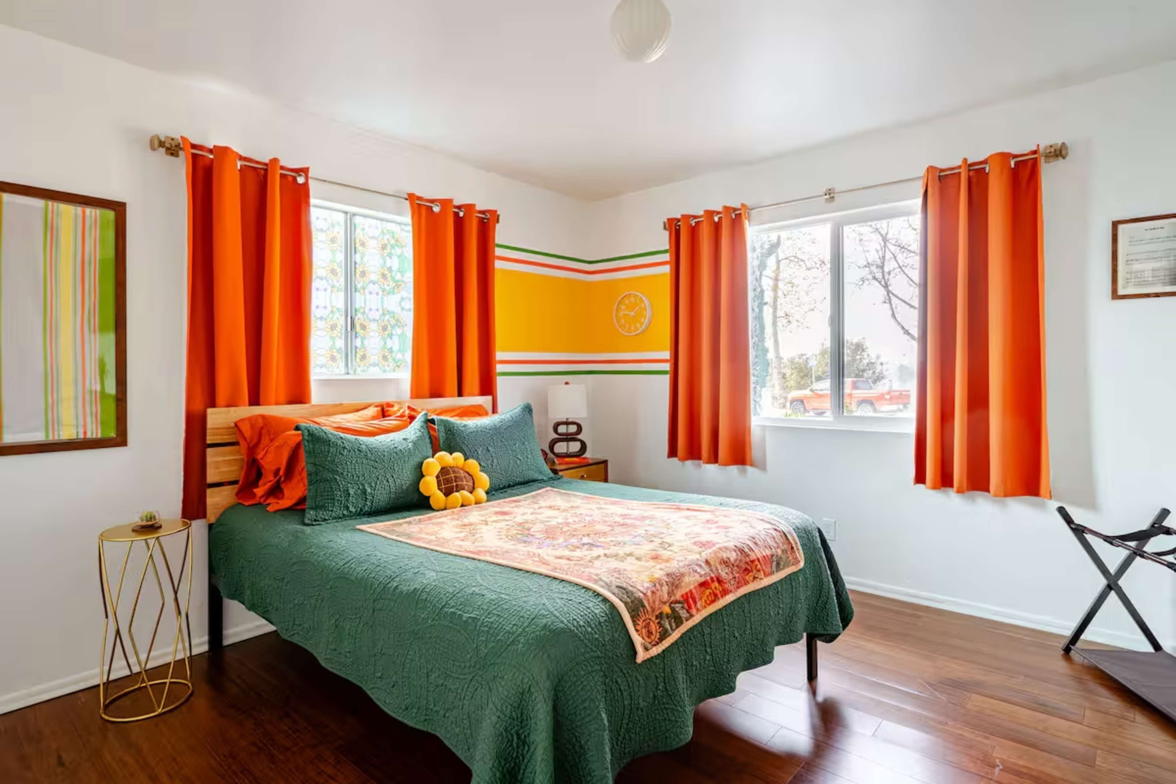 The image shows a brightly decorated bedroom featuring a green bedspread, orange curtains, and large windows.
