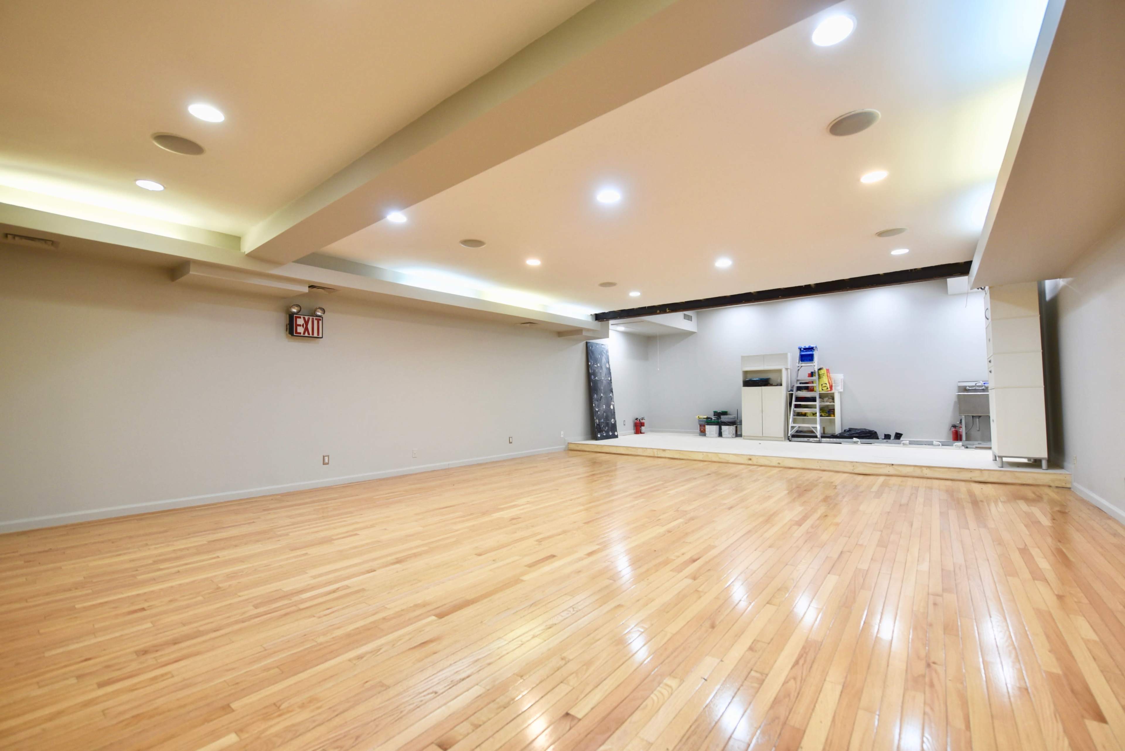 A spacious, empty room with polished hardwood floors, bright ceiling lights, and a raised platform at the far end.