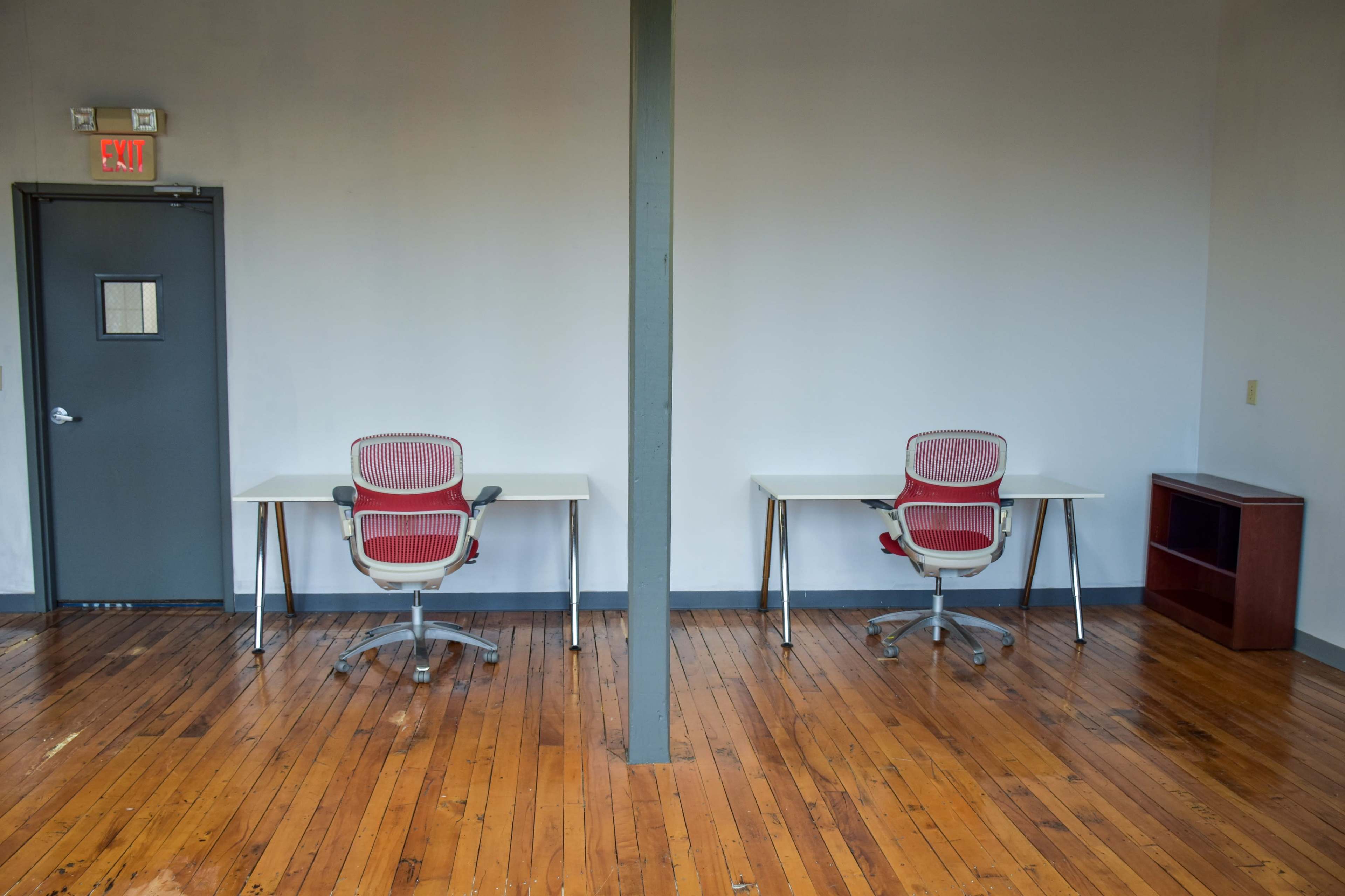 The image shows two empty desks with chairs facing each other in a room with wooden floors and a gray wall.
