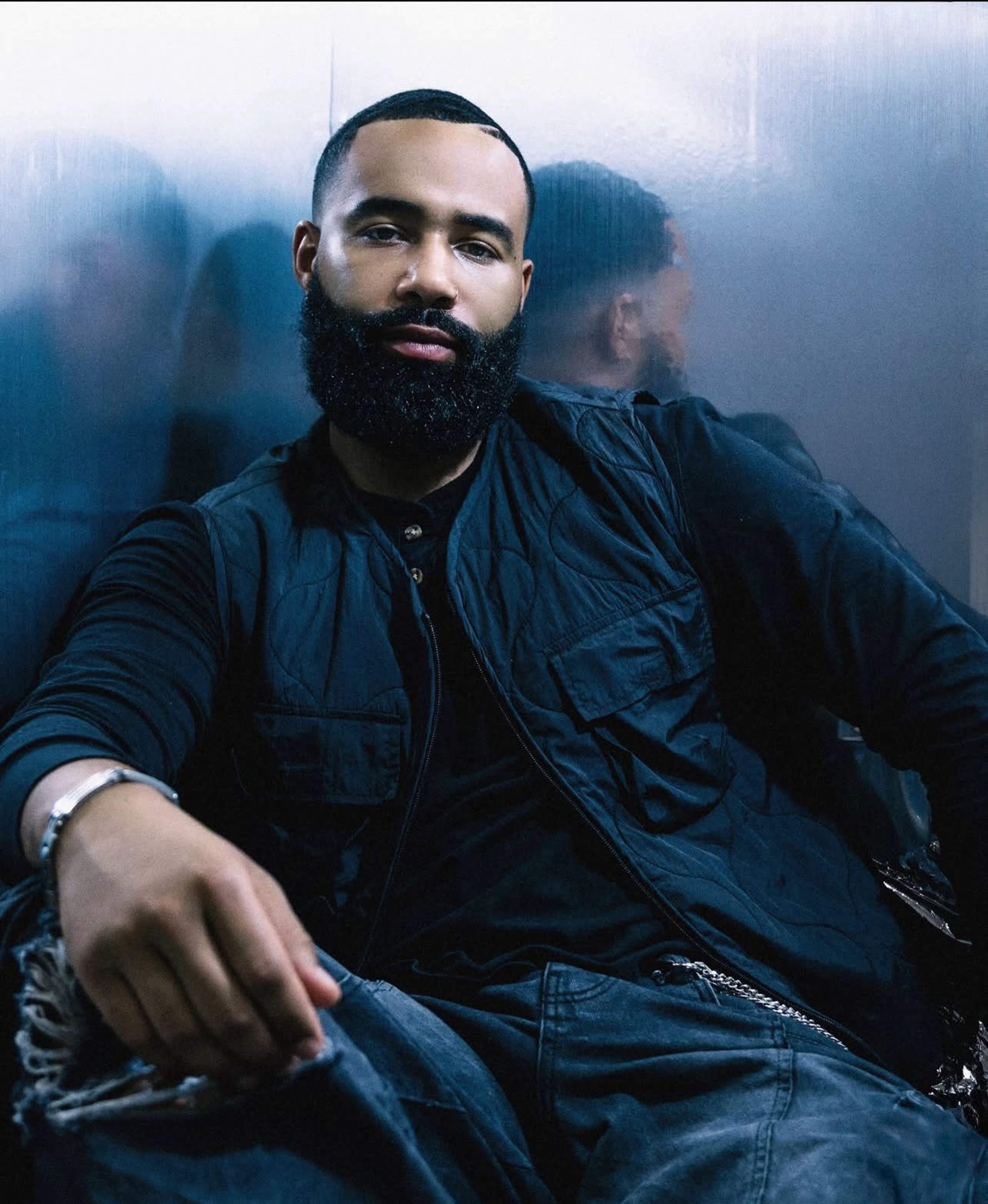 A man with a beard sits casually against a reflective surface, dressed in a black vest and loose-fitting pants.