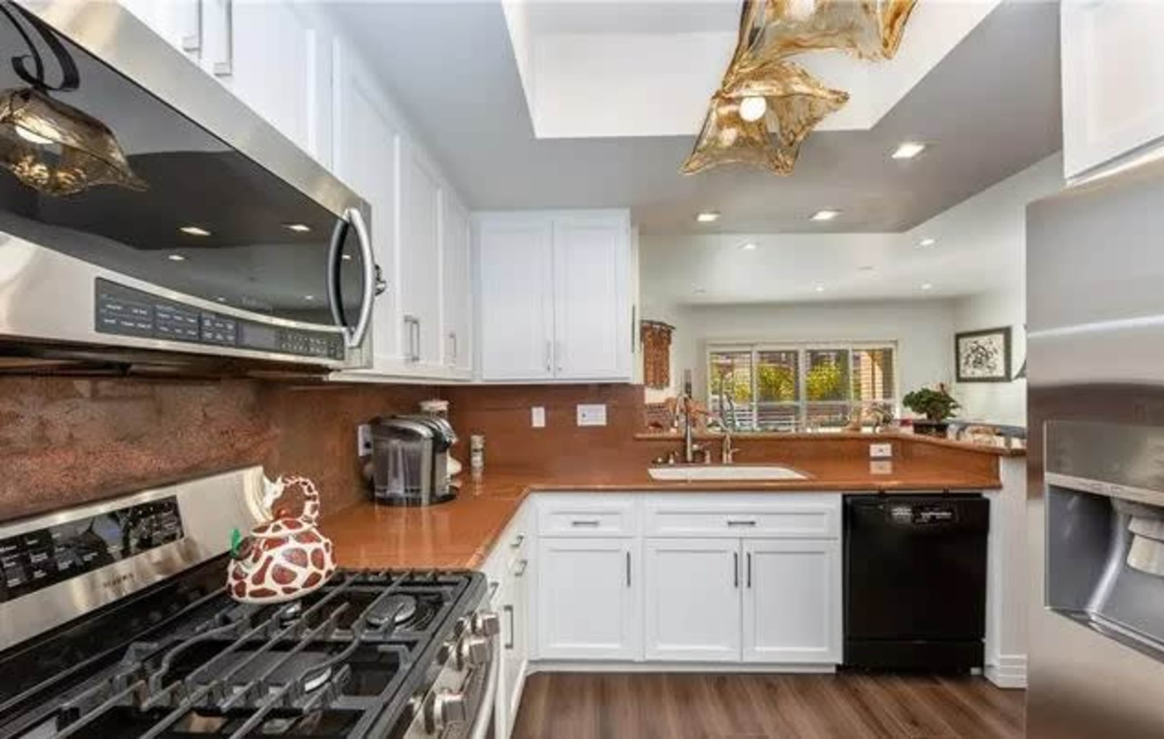 A modern kitchen features stainless steel appliances, white cabinetry, and a brown countertop with a view into a well-lit adjacent room.
