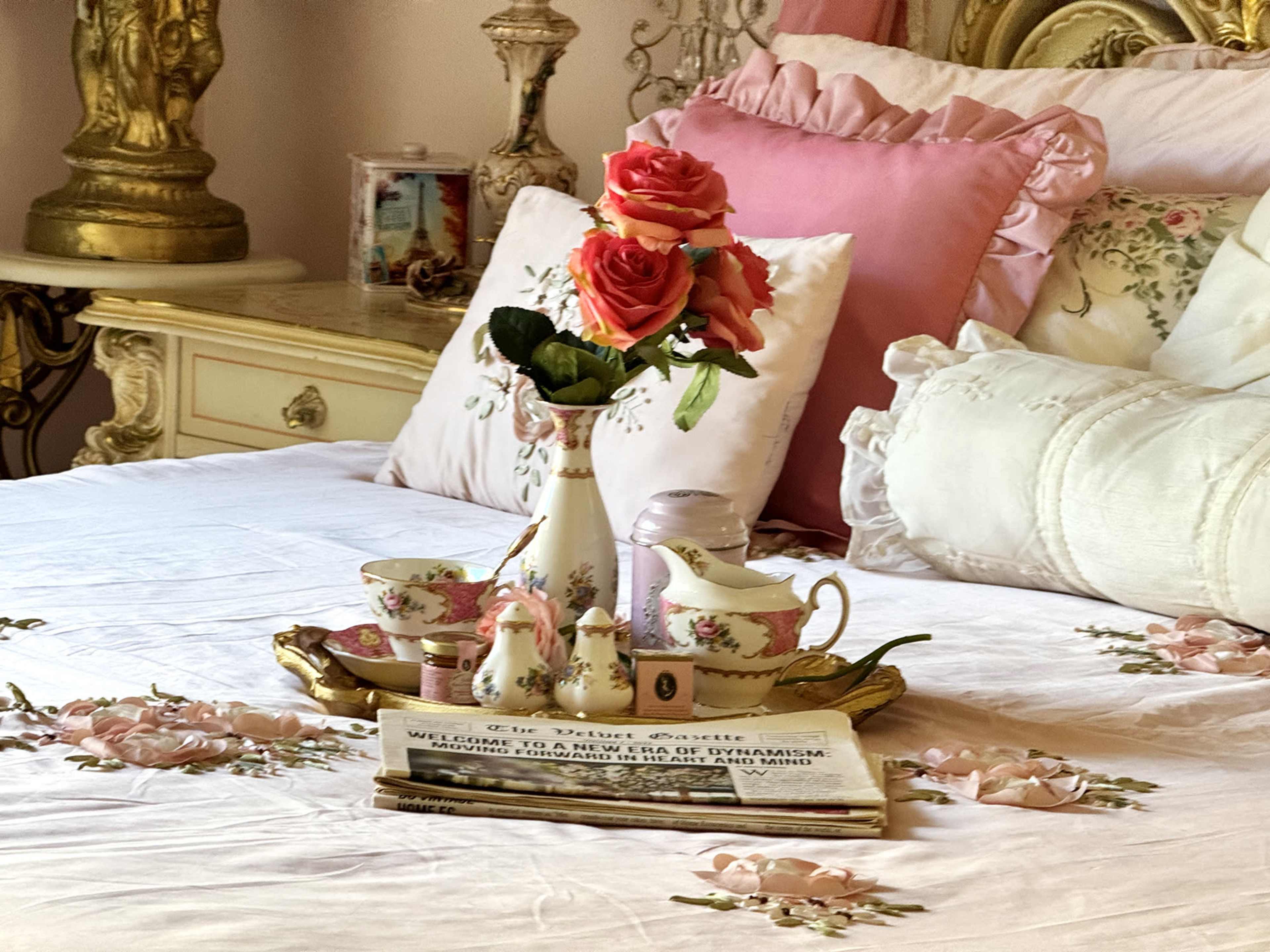 A floral arrangement of pink roses is displayed on a tray next to a tea set and a newspaper on a neatly made bed with decorative pillows.