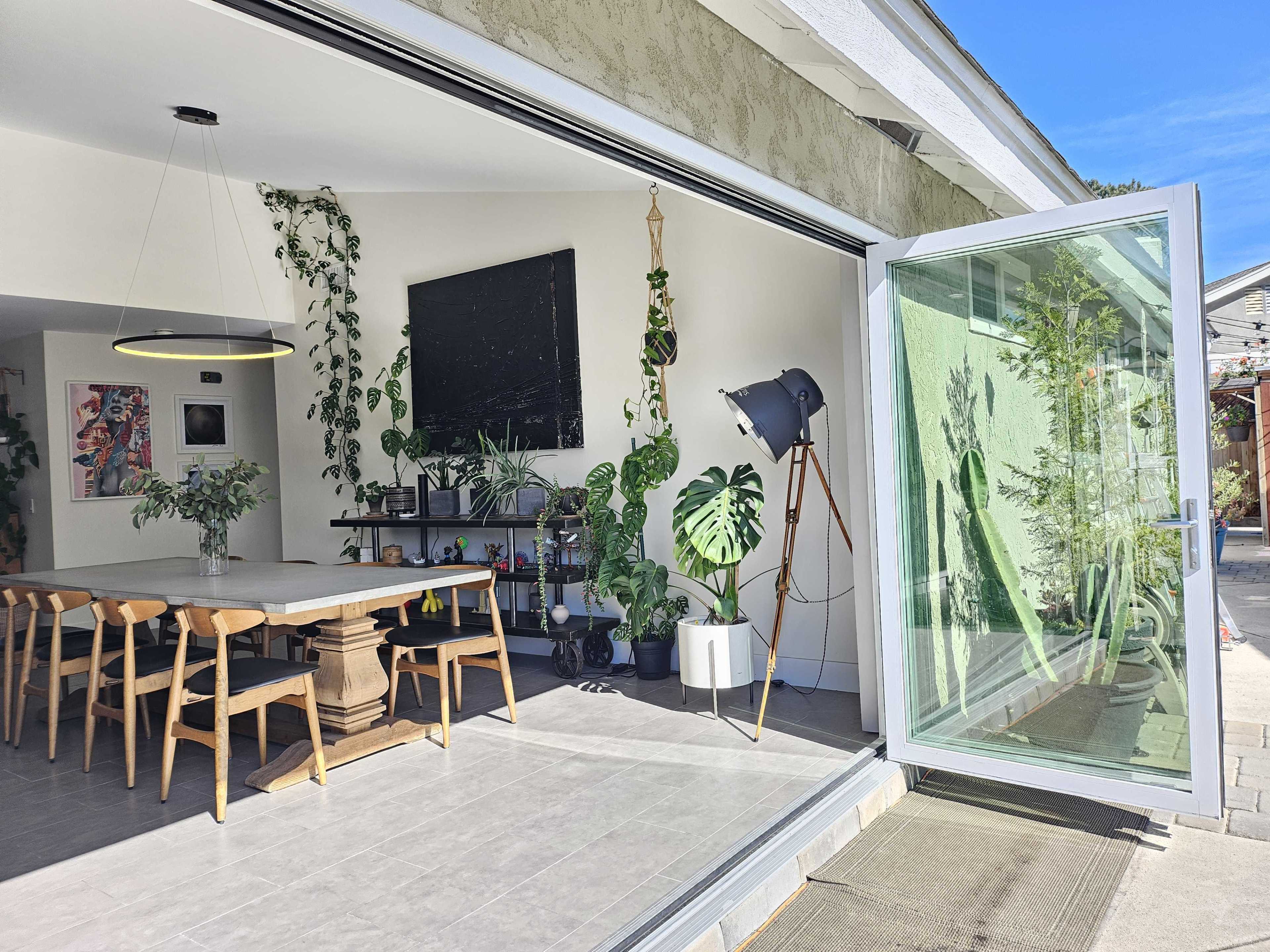 A spacious dining area features a long wooden table surrounded by chairs, large windows open to an outdoor patio, and several indoor plants.