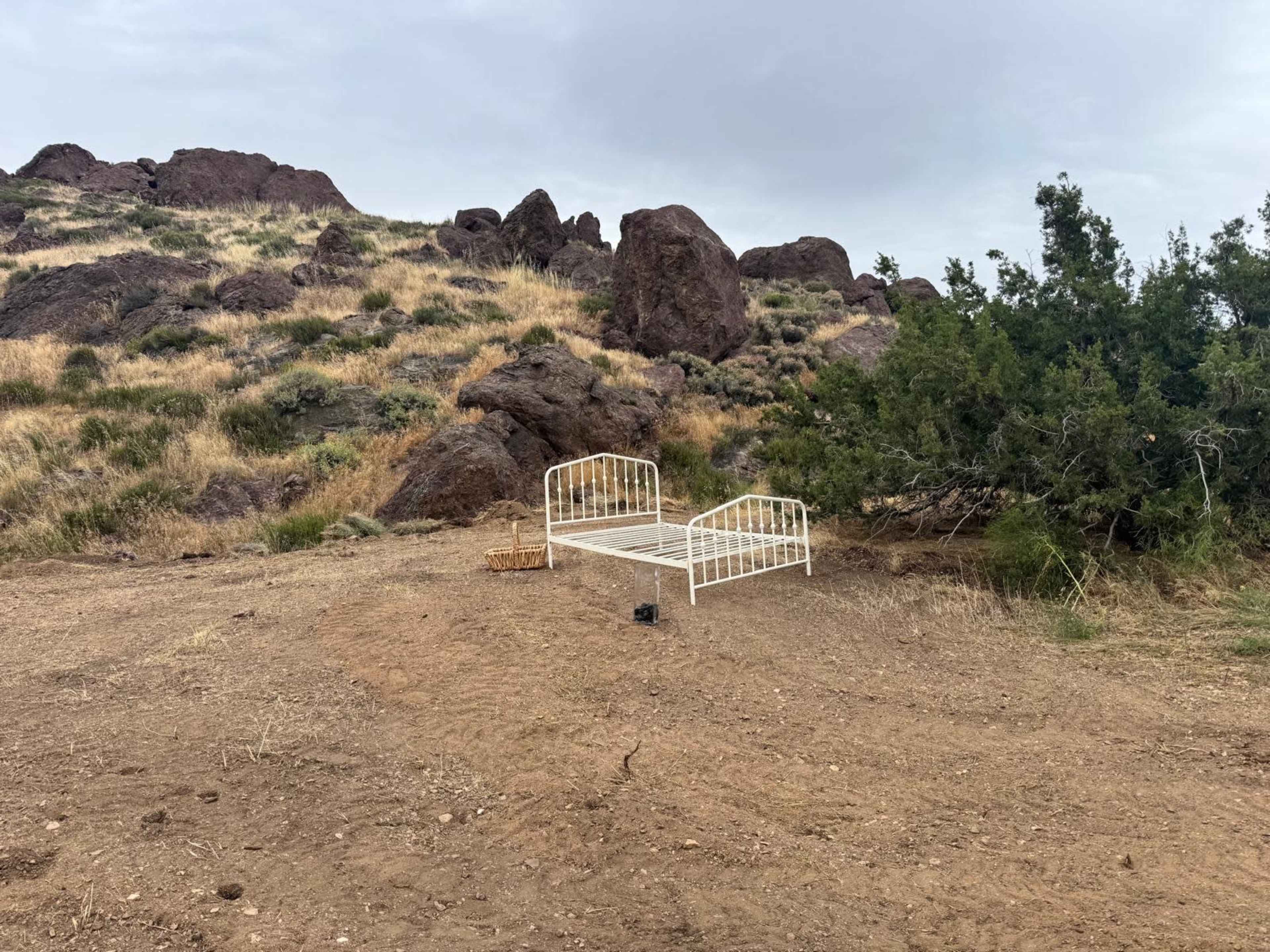 A white metal bed frame is placed on a dirt patch surrounded by rocky terrain and sparse vegetation.