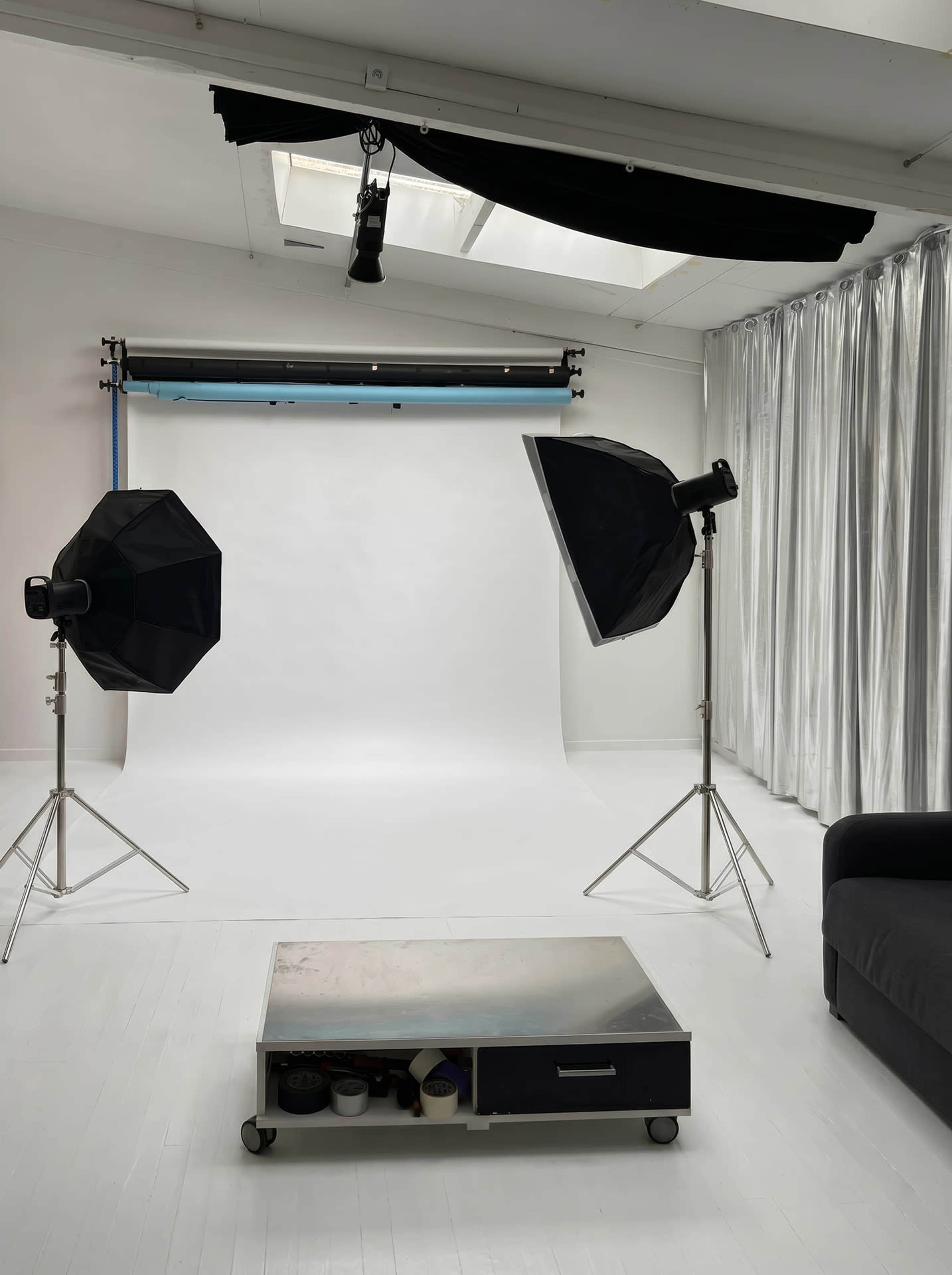 The image shows a photography studio setup with two softbox lights, a backdrop, a silver coffee table on wheels, and a sofa against a curtain.