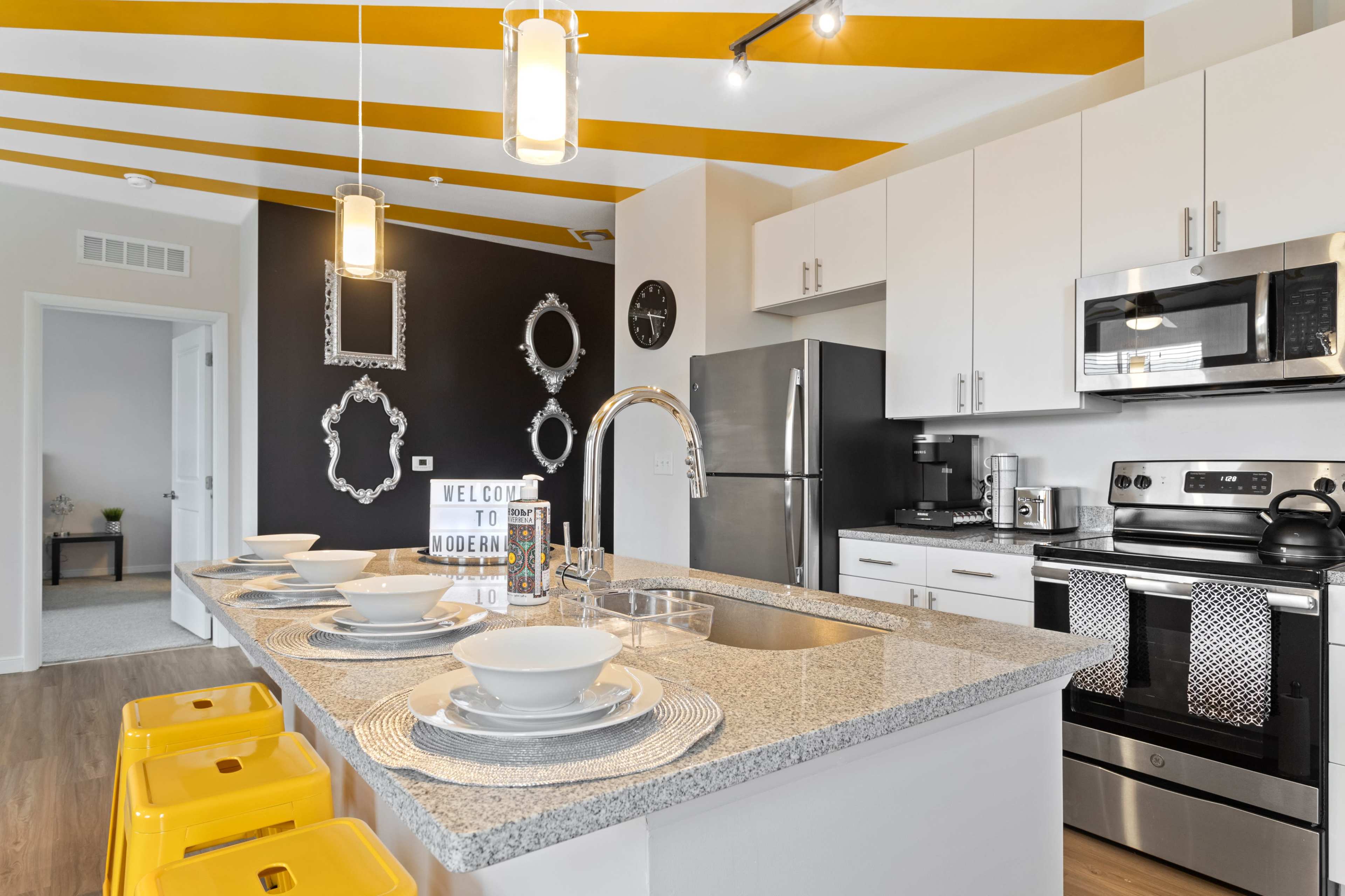 The image shows a modern kitchen with yellow-striped ceilings, gray countertops, stainless steel appliances, and a dining setup featuring white plates and yellow stools.