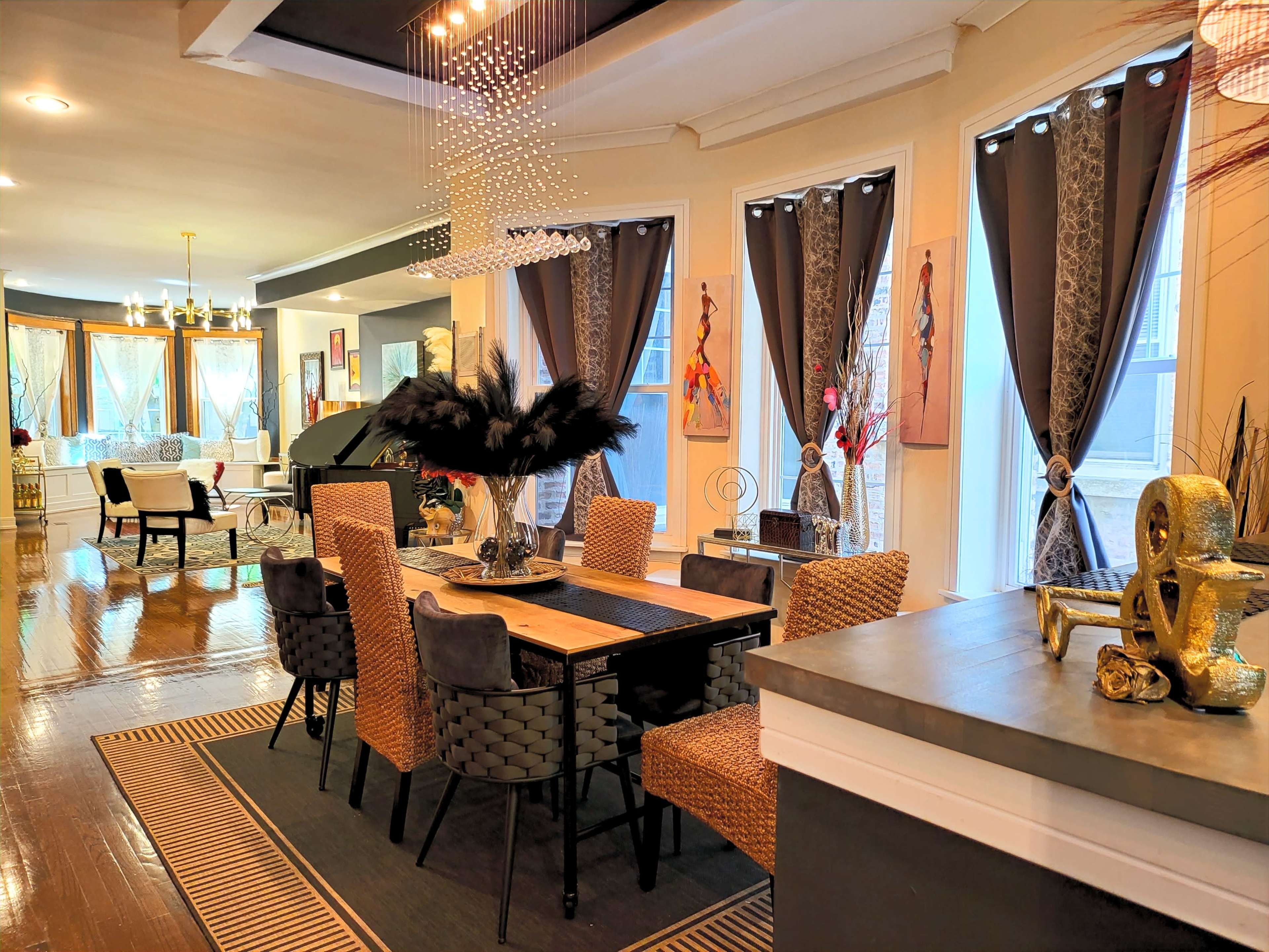 A modern dining area with a long table surrounded by woven chairs, situated in a well-lit room featuring large windows with sheer drapes.