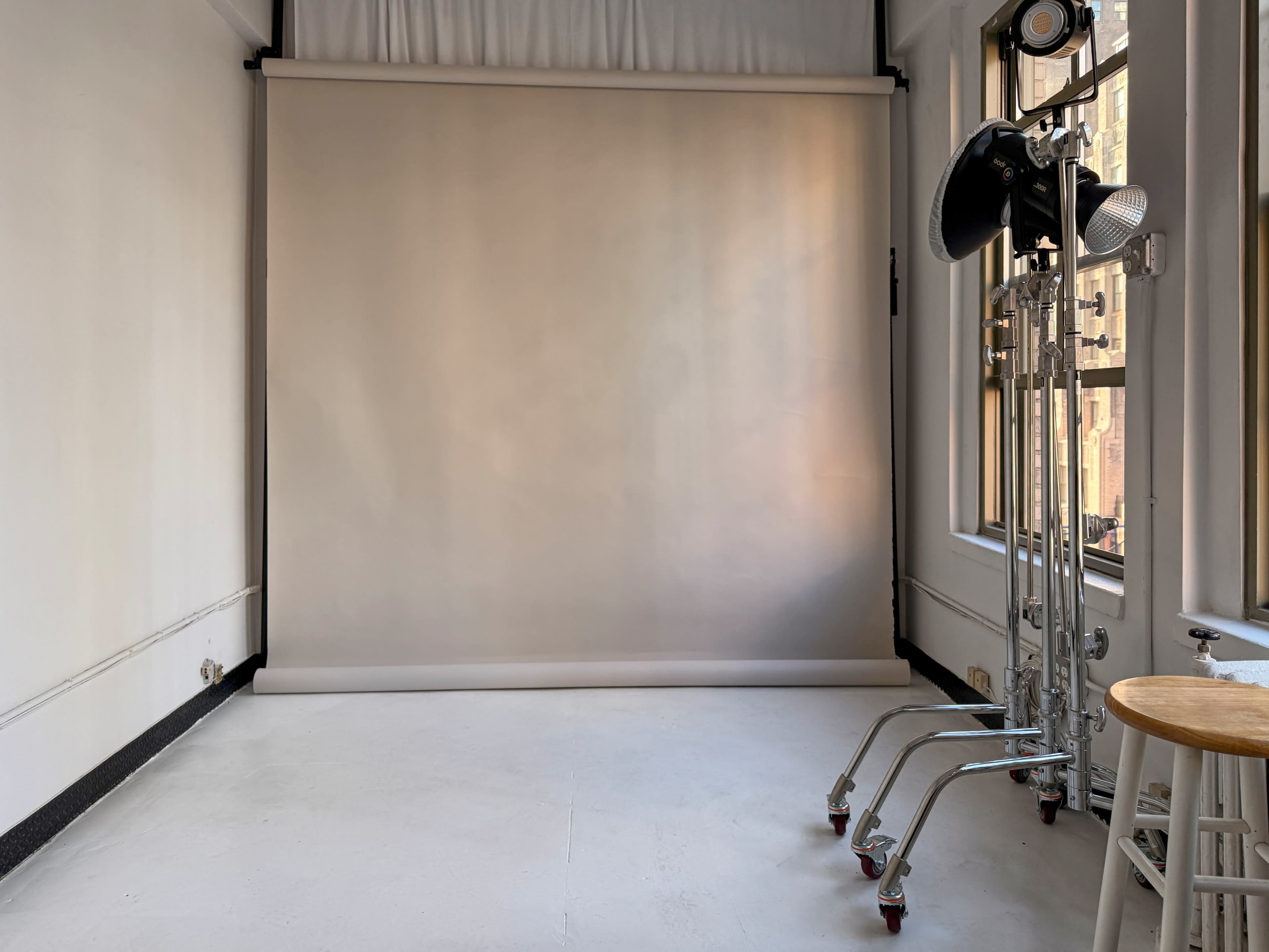 The image shows a photography studio featuring a neutral backdrop, a light stand, and a wooden stool against white walls.