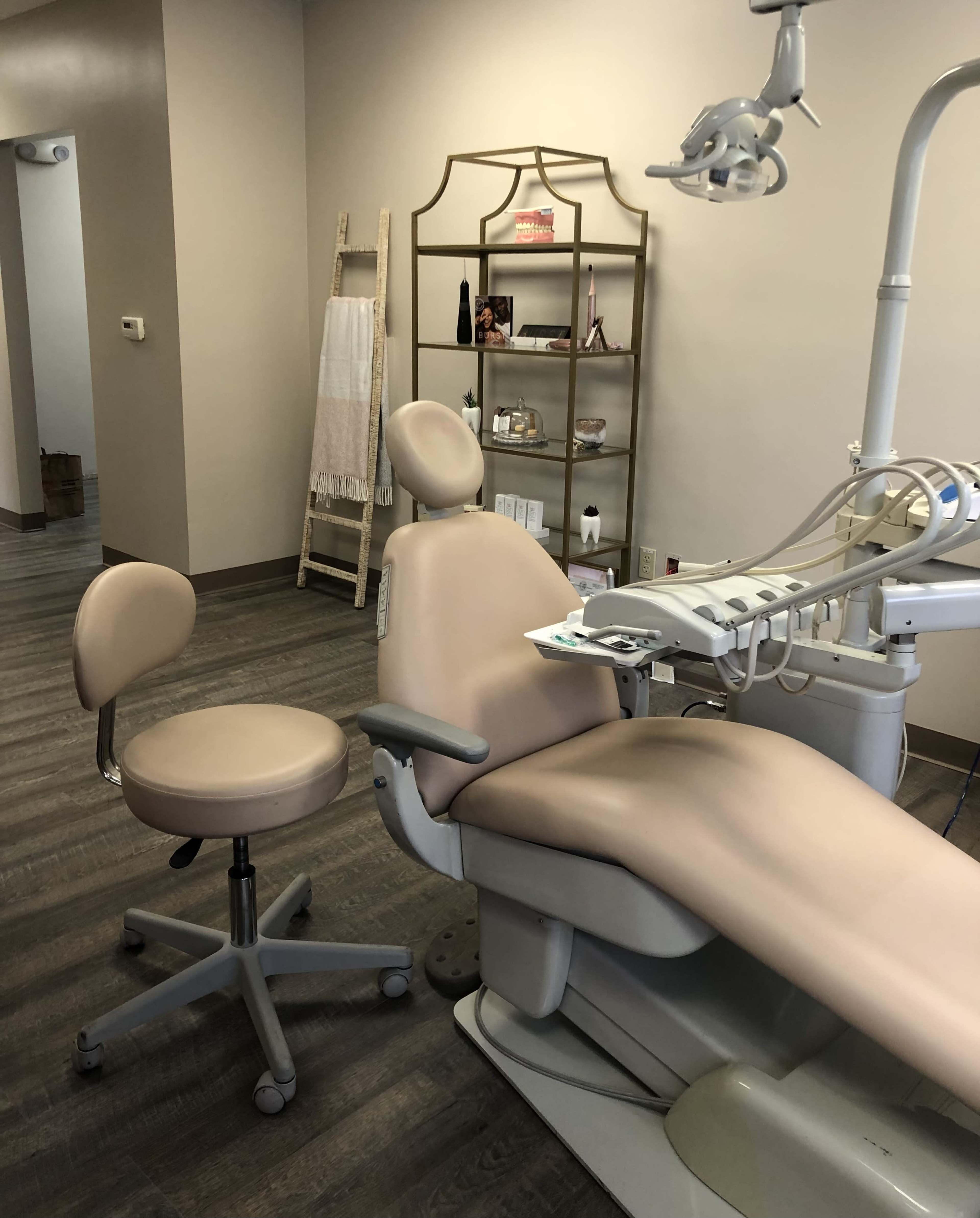 A dental examination room with a dental chair and a rolling stool, along with a shelf displaying decorative items.